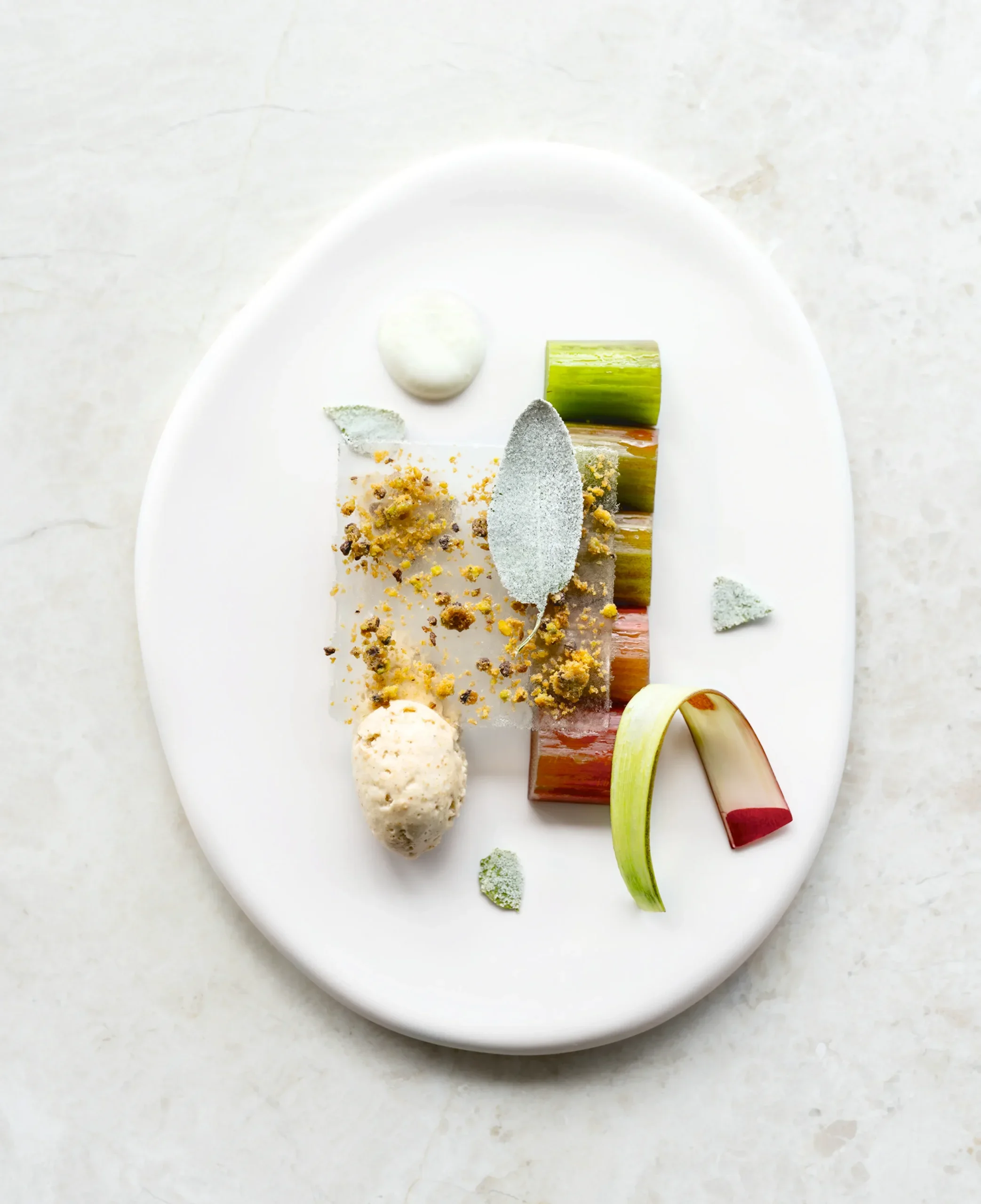 Présentation élégante d'une assiette de dessert avec diverses garnitures, comprenant un sorbet ivoire, des rouleaux de légumes verts et rouges, des morceaux croquants, et des feuilles décoratives.