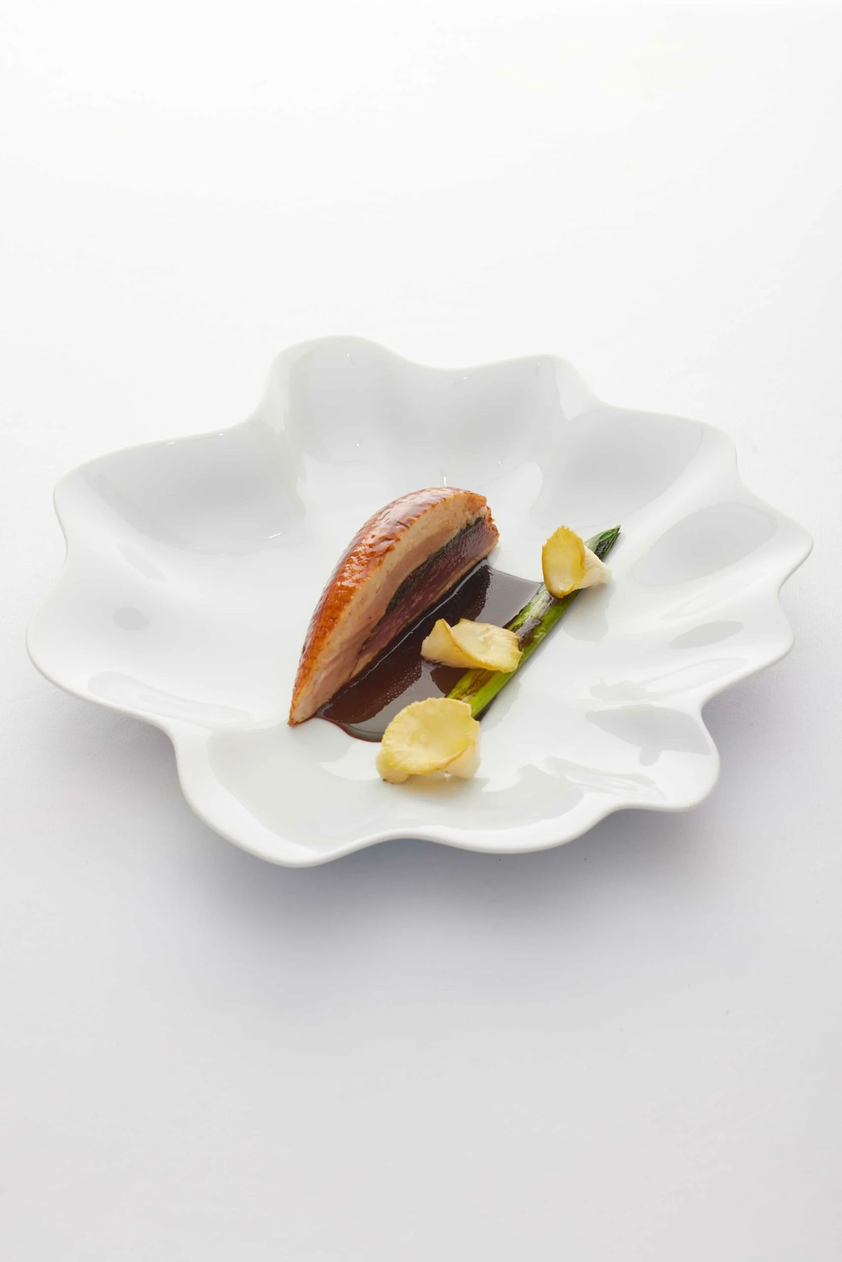 Plat de cuisine moderne avec une portion de viande, une sauce sombre, un asperge et des tranches d'ail sur une assiette blanche.