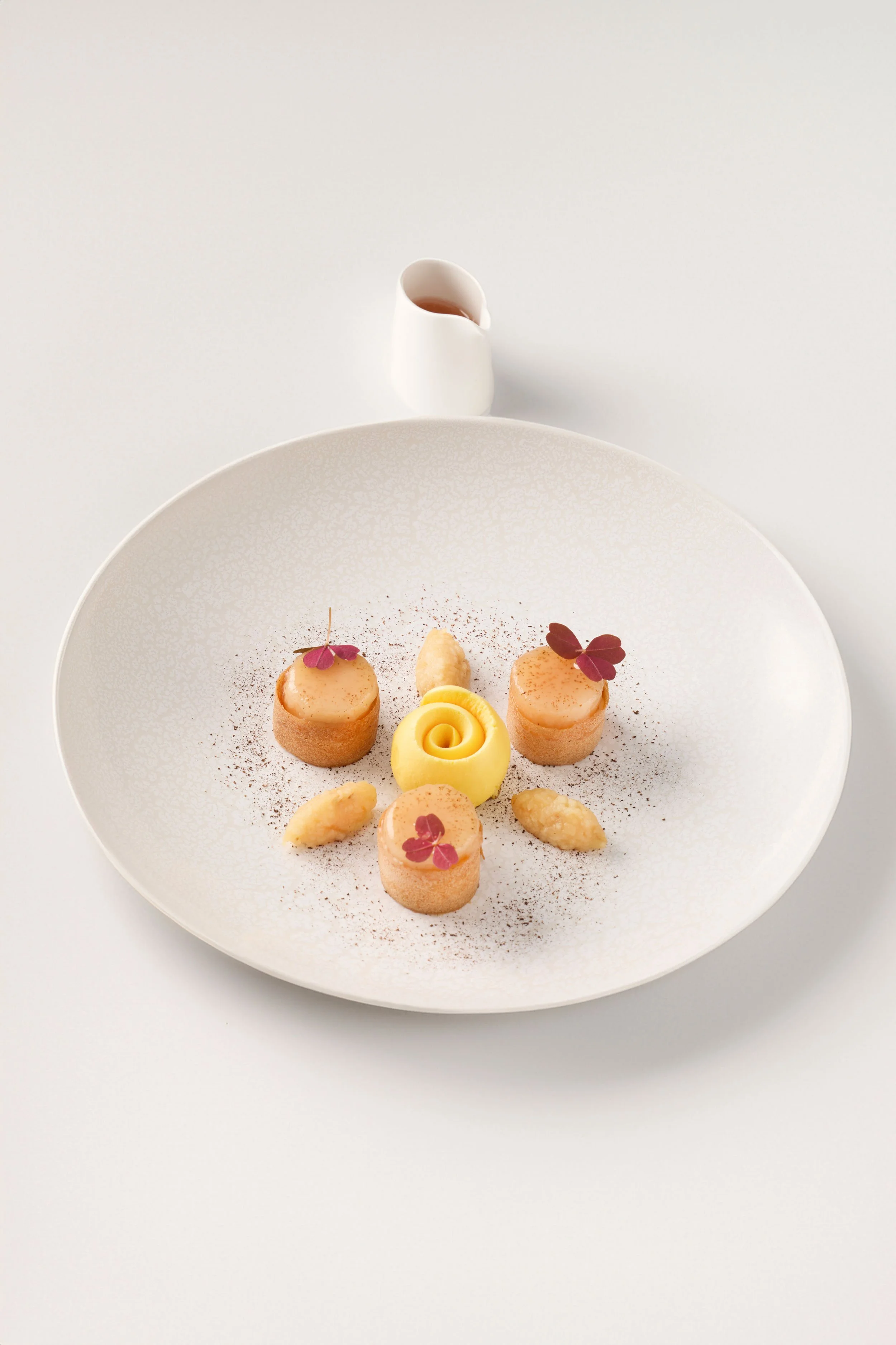 Dessert élégant présenté sur une assiette blanche avec petits éléments décoratifs et une petite cruche blanche contenant une sauce.