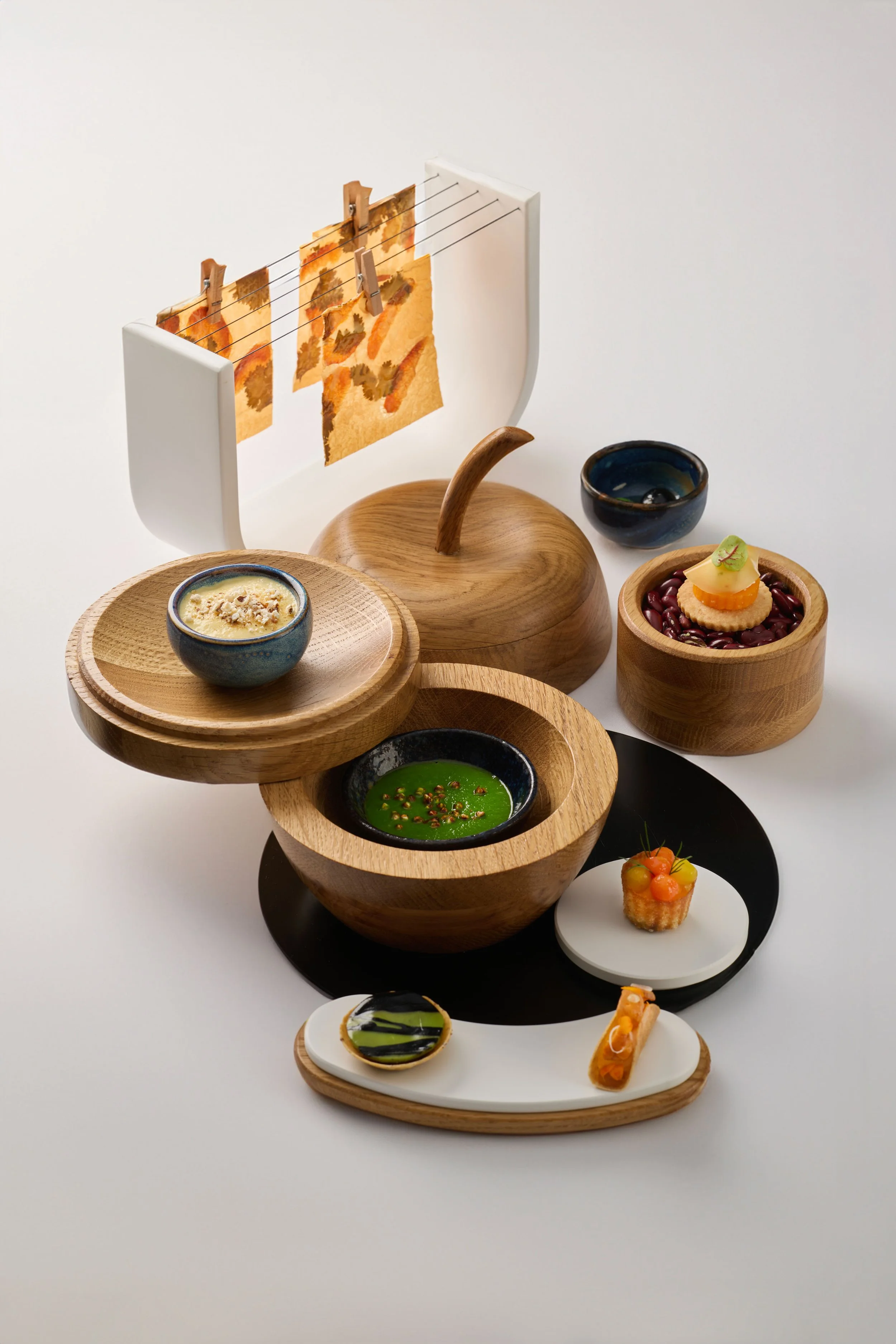 Une présentation de plusieurs petits plats et amuse-bouches dans différents bols et assiettes en bois, avec un dispositif suspendu à clips et fils à l'arrière.
