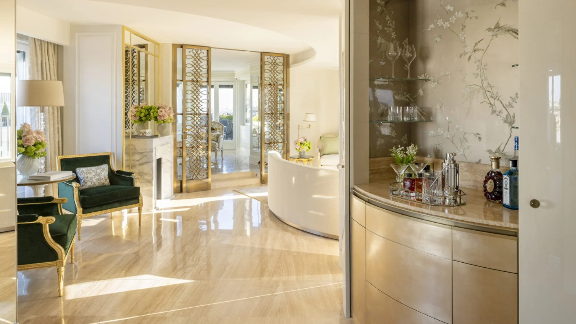 Intérieur d'un salon lumineux avec des fauteuils verts, fleurs en vase, séparateurs en métal doré, et une vue sur un balcon ou une terrasse.