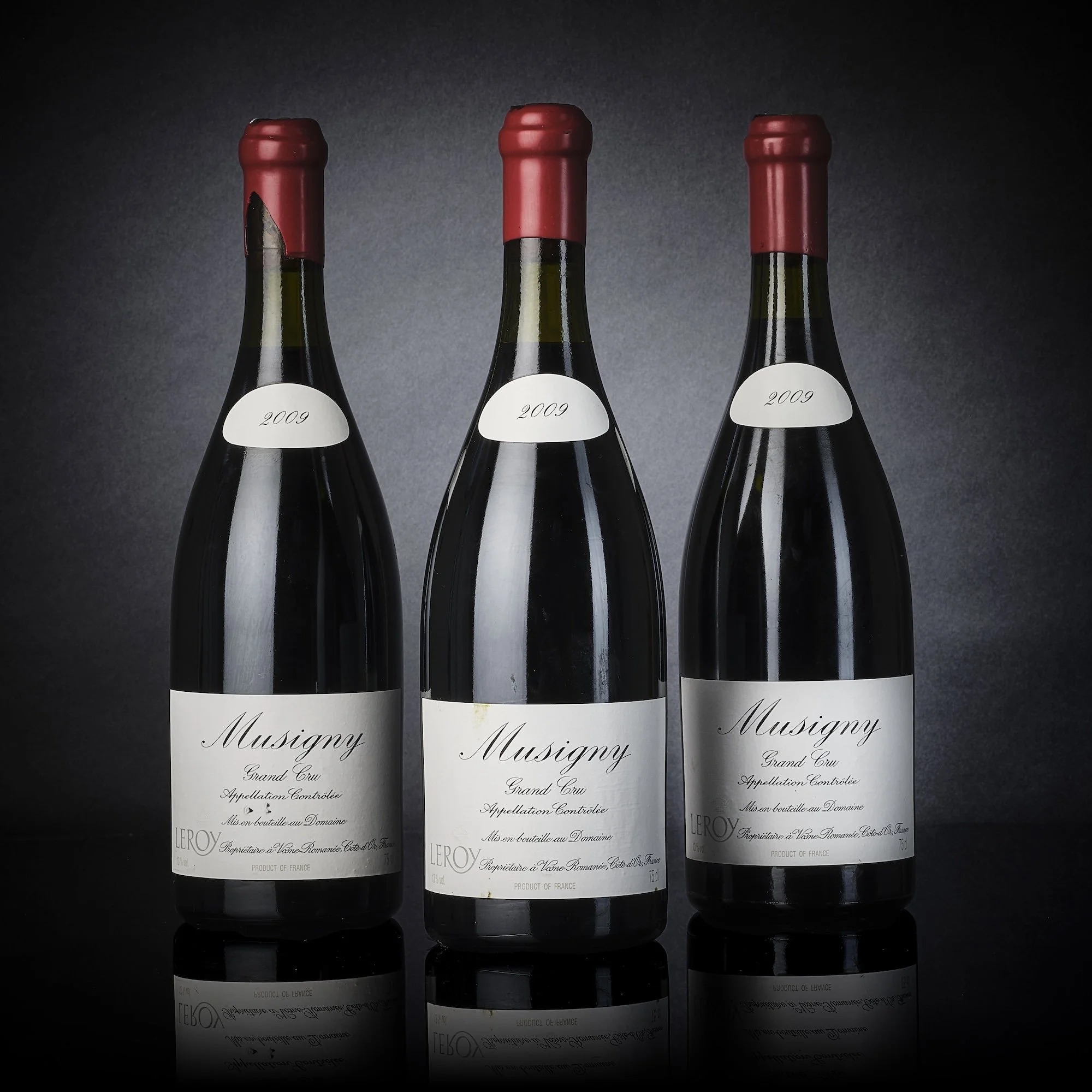 Trois bouteilles de vin rouge avec étiquettes noires et blanches, de la marque Musigny, année 2009, positionnées côte à côte sur fond noir, avec leur reflet reflété sous elles.