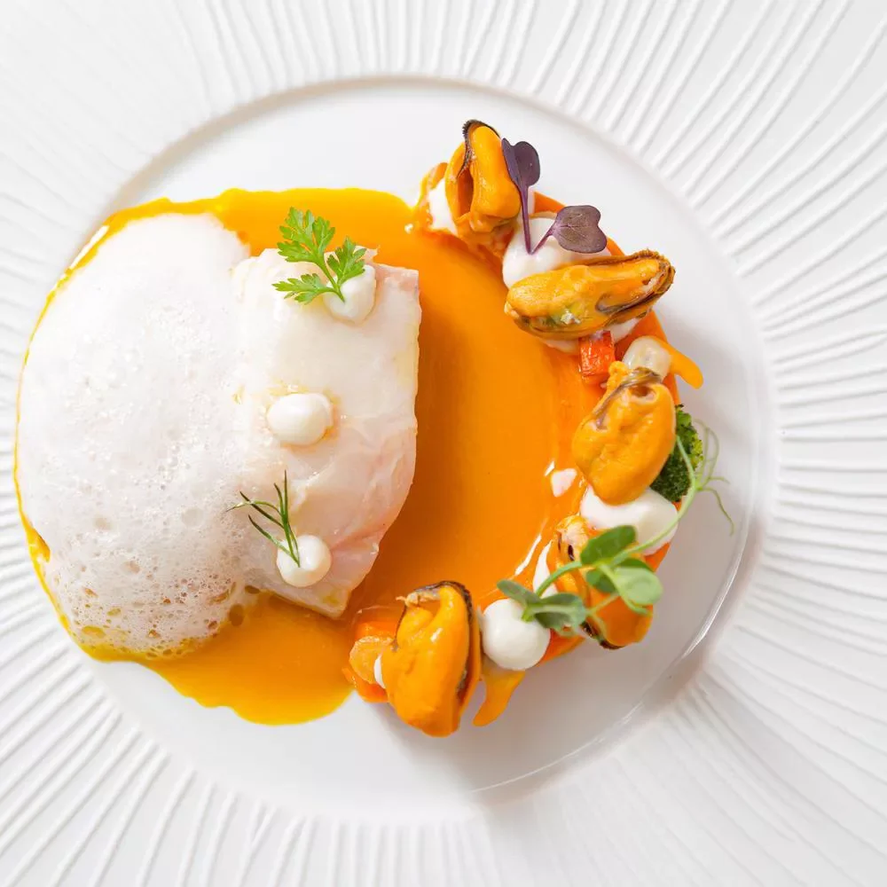 Assiette de fruits de mer avec mollusques, sauce orange, mousse blanche, microgreens et décor végétal sur une assiette blanche