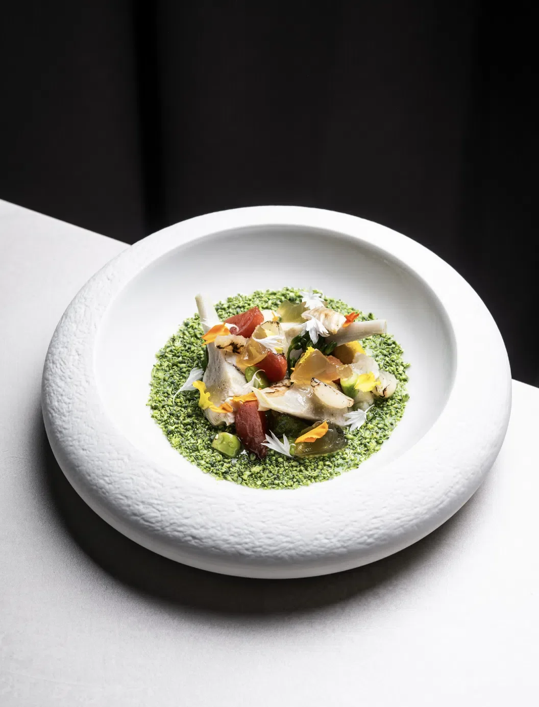 Une assiette de présentation moderne avec une garniture de légumes, de fleurs comestibles, et de petits morceaux de poisson ou de viande, entourée d'une fine couche de mousse ou de purée verte.