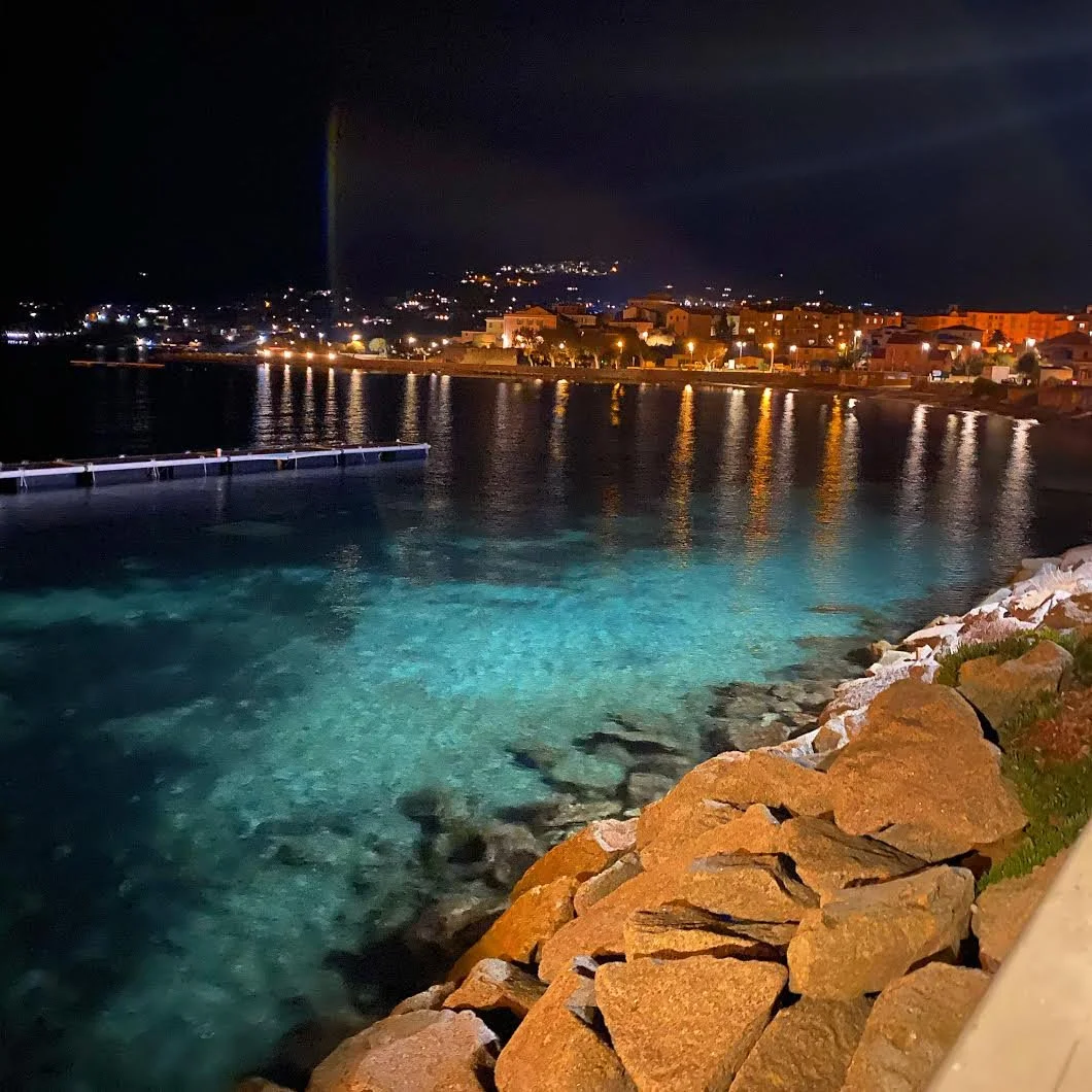 Vue nocturne d'une côte avec des bâtiments illuminés, des rochers au premier plan et un reflet des lumières sur l'eau.