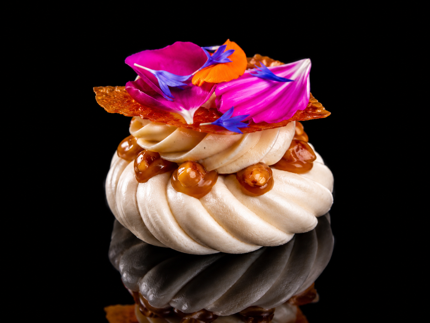 Un dessert sophistiqué composé de cerises caramelisées, crème fouettée, pétales de fleurs violettes, roses, orange et feuilles comestibles, avec une décoration florale colorée, présenté sur un fond noir.