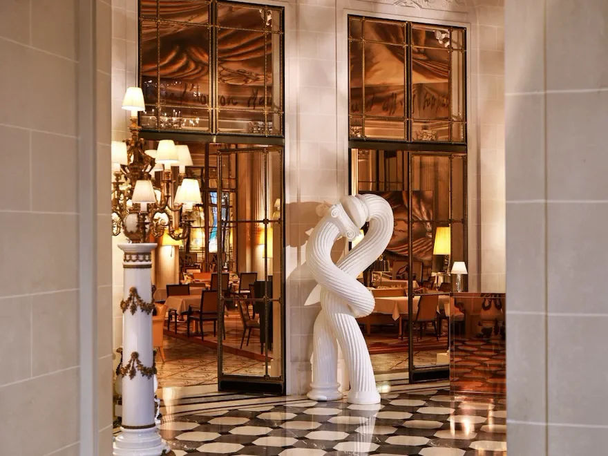 Intérieur d'un restaurant ou d'un hôtel élégant avec une sculpture de deux oiseaux en blanc au centre. La pièce est ornée de miroirs, d'une colonne décorative avec des lampes, et dispose d'une salle à manger avec des tables et des chaises.
