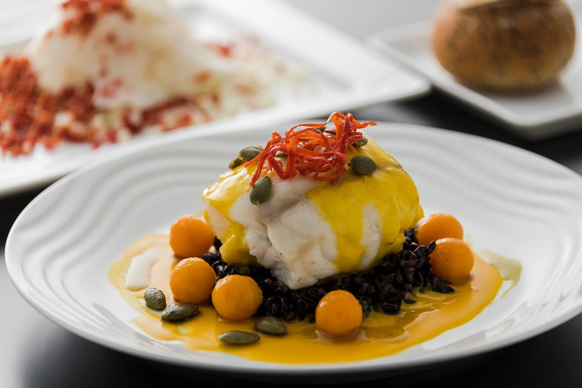 Plat de poisson blanc avec sauce jaune, entouré de billes orange et de graines, décoré de julienne de poivron rouge. Arrière-plan flou avec autres plats.