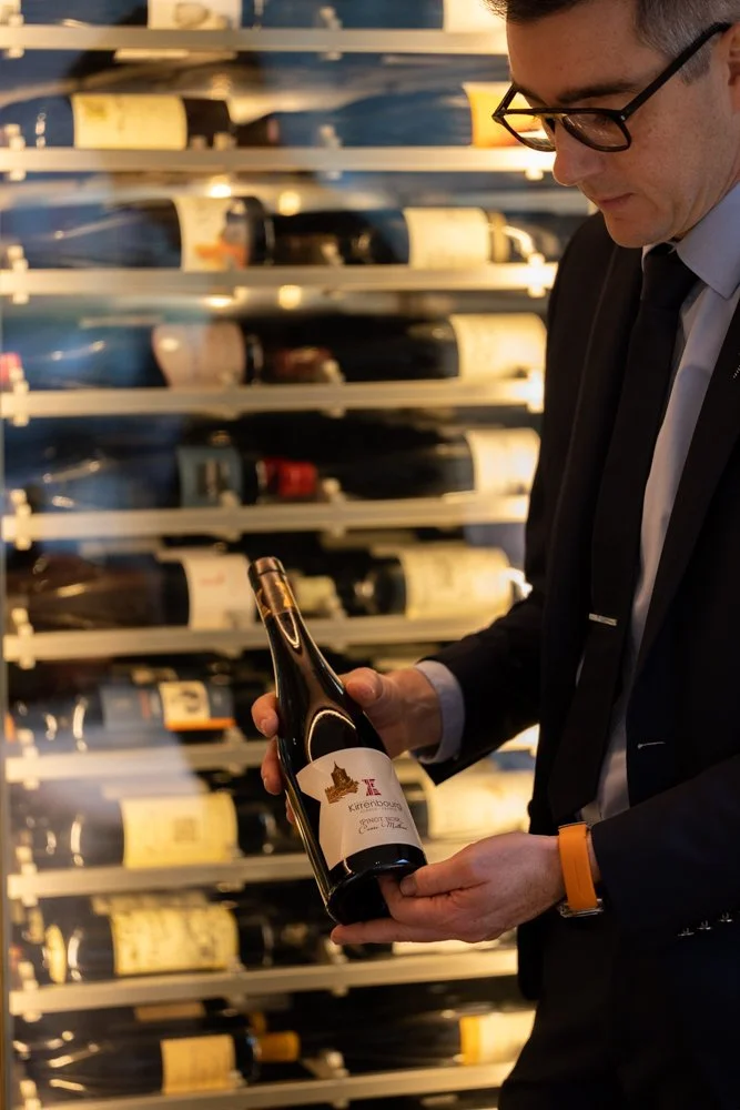 Homme Habillé en costume, portant des lunettes, regardant une bouteille de vin dans un magasin de vins.