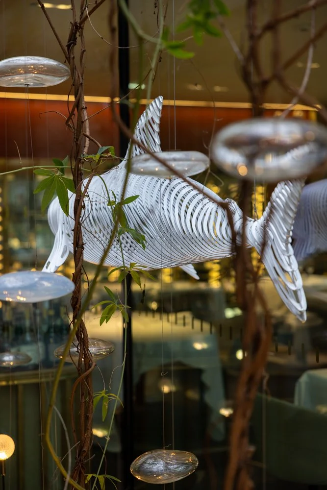 Une décoration suspendue représentant un poisson en métal, entourée de branches de vigne et de petites élytres en verre dans un intérieur moderne.
