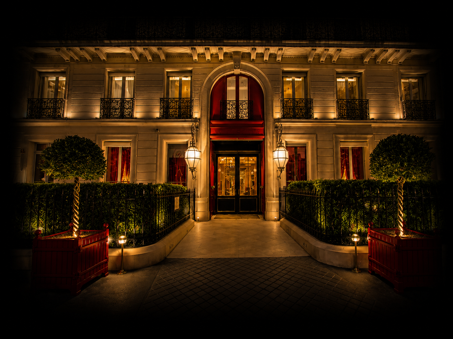 Façade éclairée d'un bâtiment élégant avec fenêtres à volets, portico, lampes extérieures et espace vert décoratif.