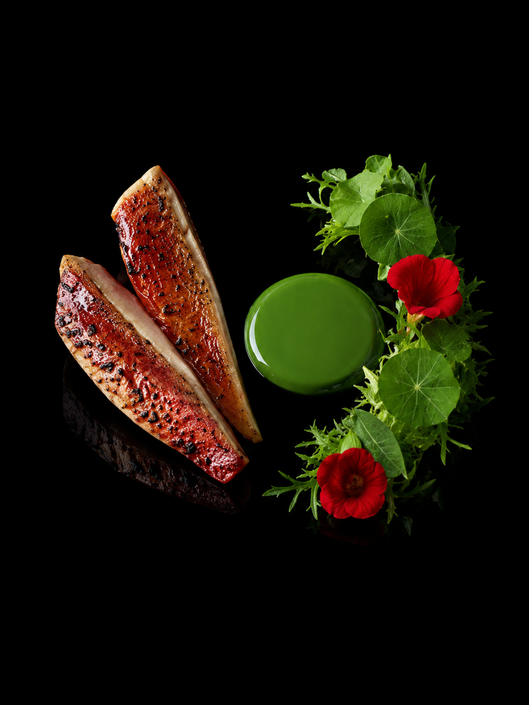 Deux tranches de poisson grillé à la surface orange et noir et un verre de jus vert, entourés de feuilles vertes et de fleurs rouges, sur fond noir.
