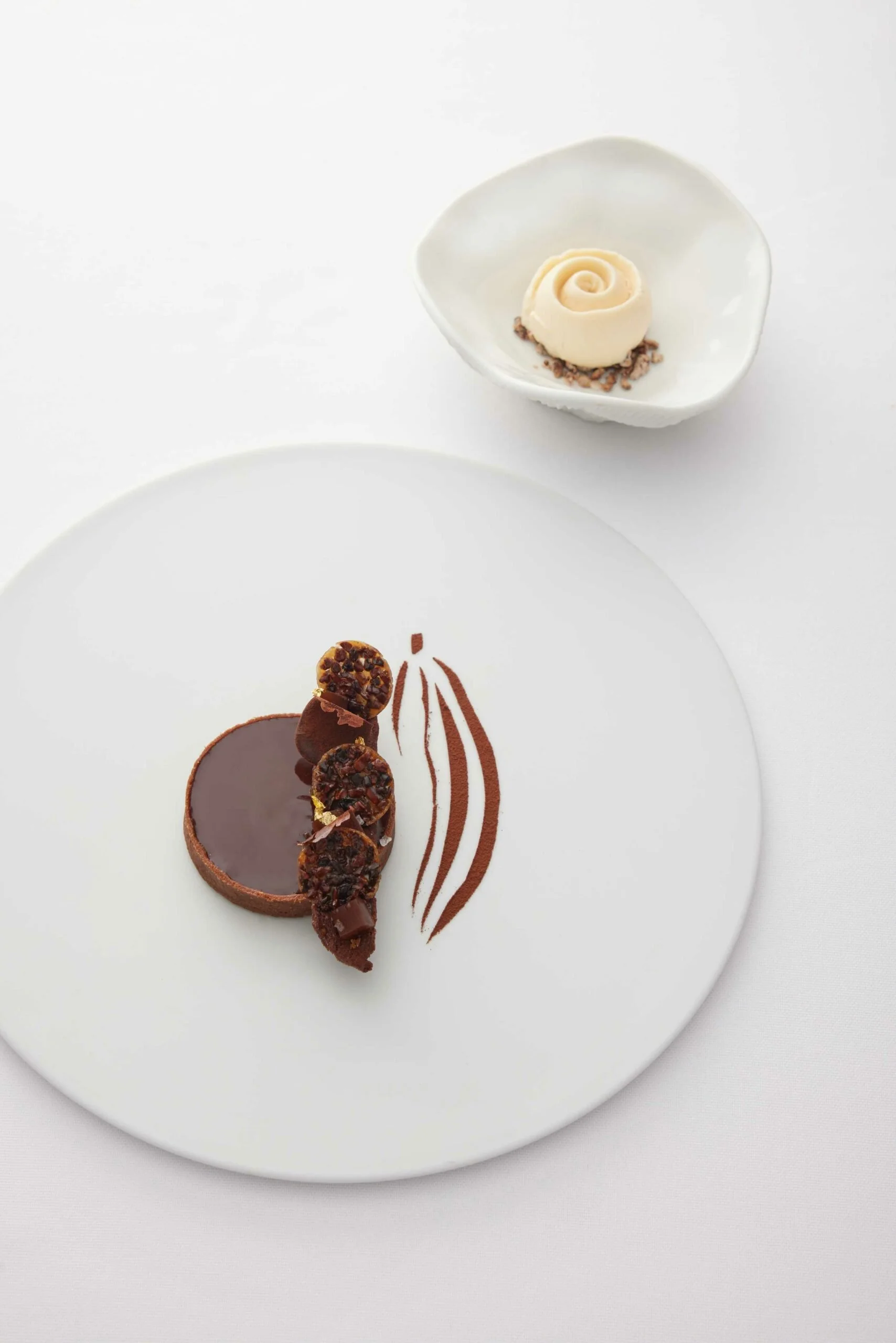 Dessert chocolaté avec trois petits entremets, décoré de pâte à tartiner et de cacao en poudre, présenté sur une assiette blanche avec un dessin en cacao.