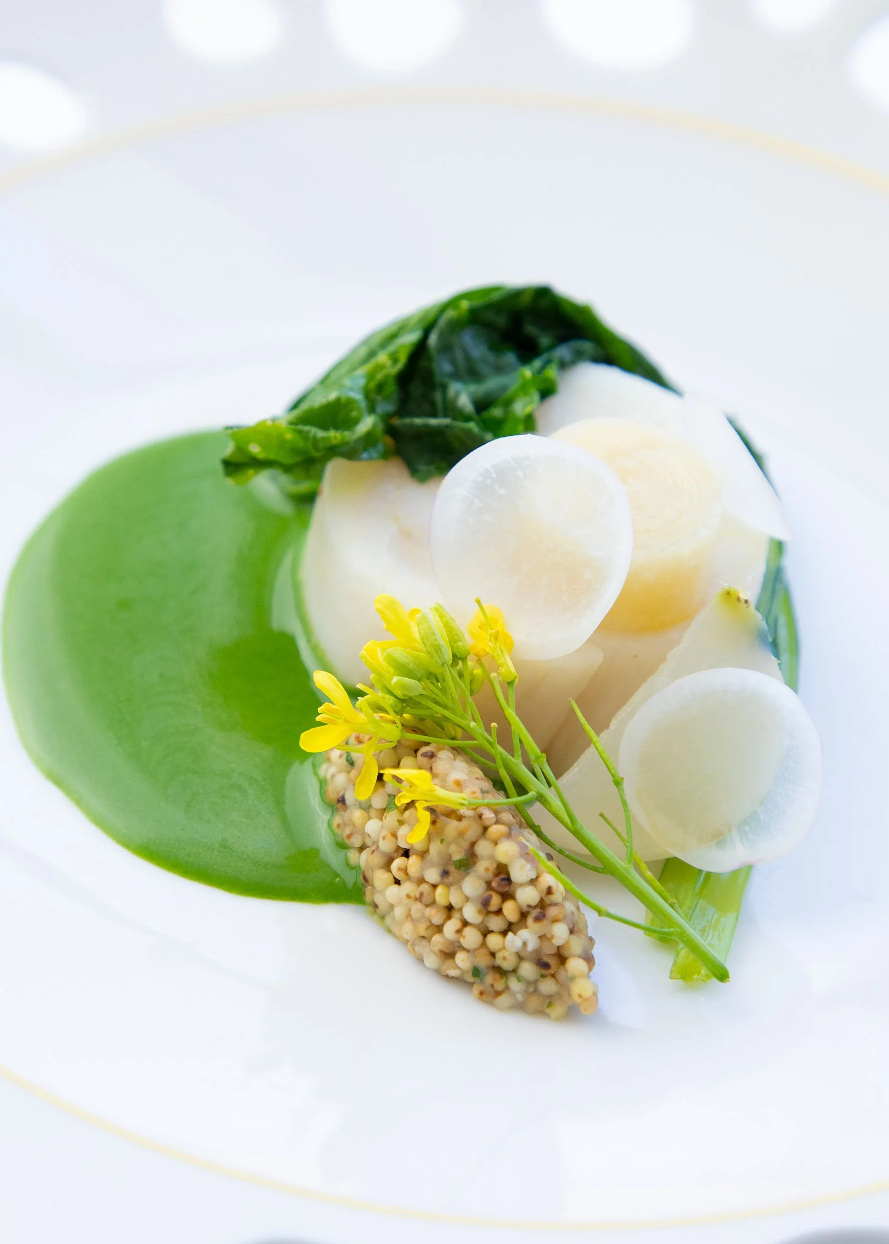 Assiette de cuisine végétarienne avec purée verte, pousses, légumes blancs, graines de moutarde, et fleurs comestibles.