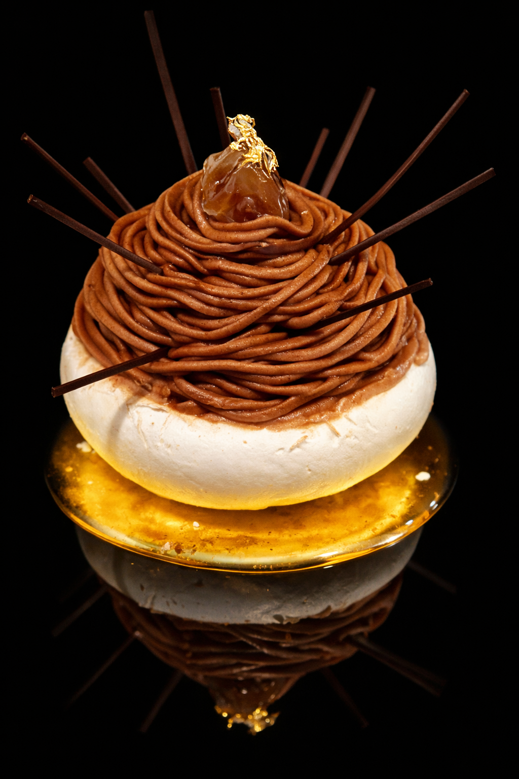 Un dessert sophistiqué avec une base de meringue blanche surmontée de vermicelles de chocolat en spirale, décoré avec une perle de caramel ou de ganache et des bâtonnets de chocolat, présenté sur une surface noire.