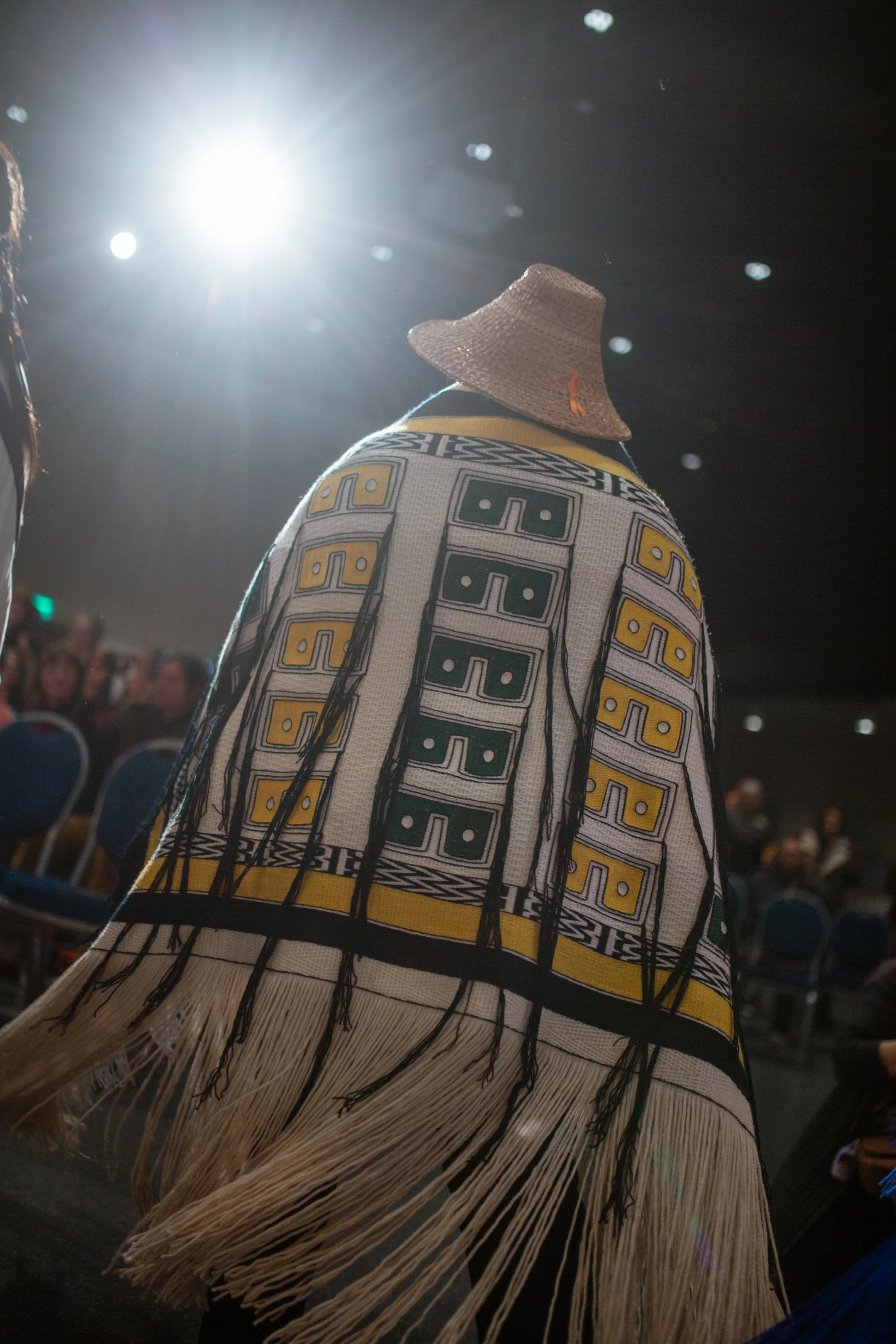 A person dressed in a colorful Tlingit Ravenstail robe and straw hat standing at a cultural event, with an audience visible in the background and bright light overhead.