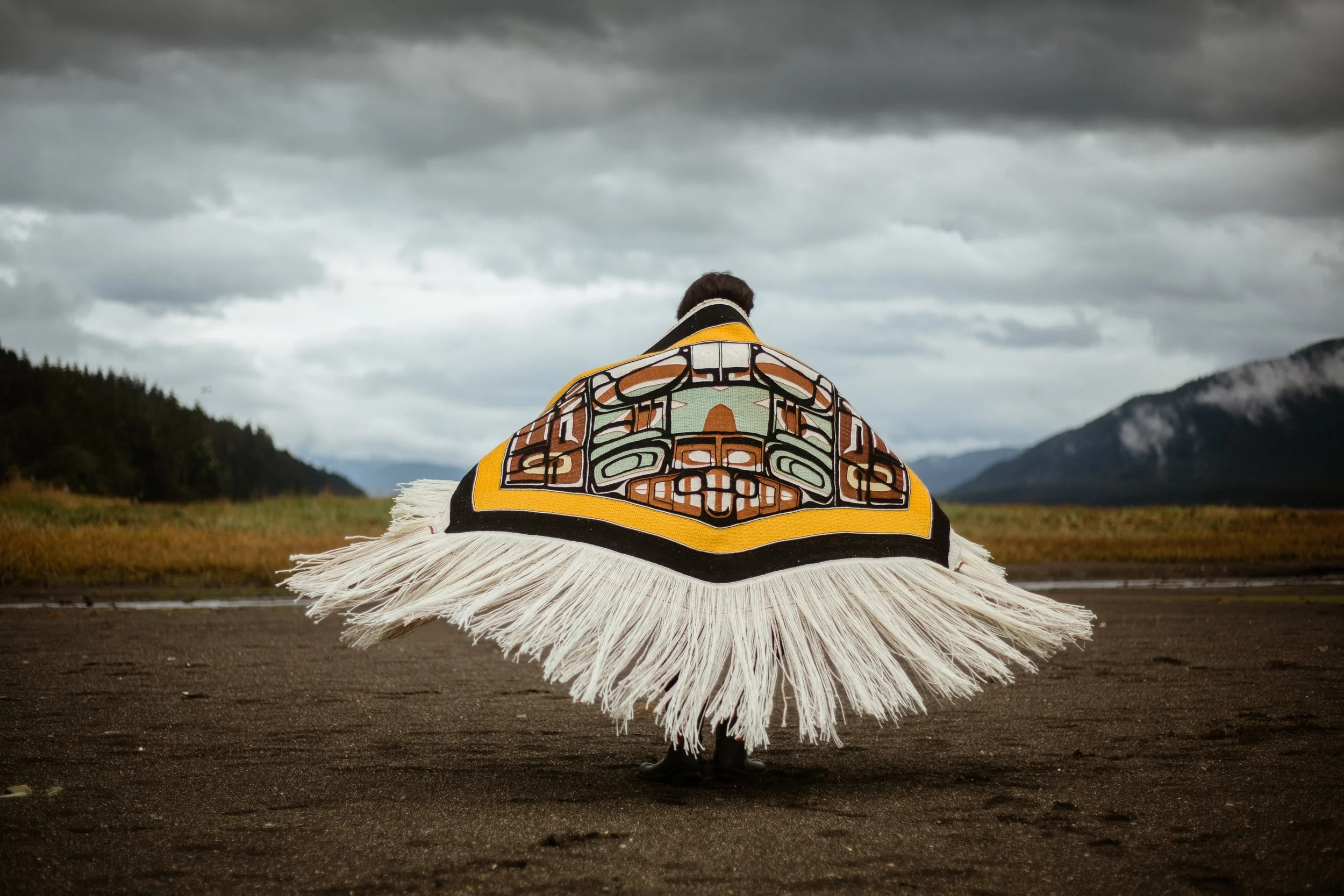 A person wearing a traditional Tlingit Chilkat woven blanket standing on a dark Southeast Alaska beach, facing away toward distant mountains and cloudy sky.