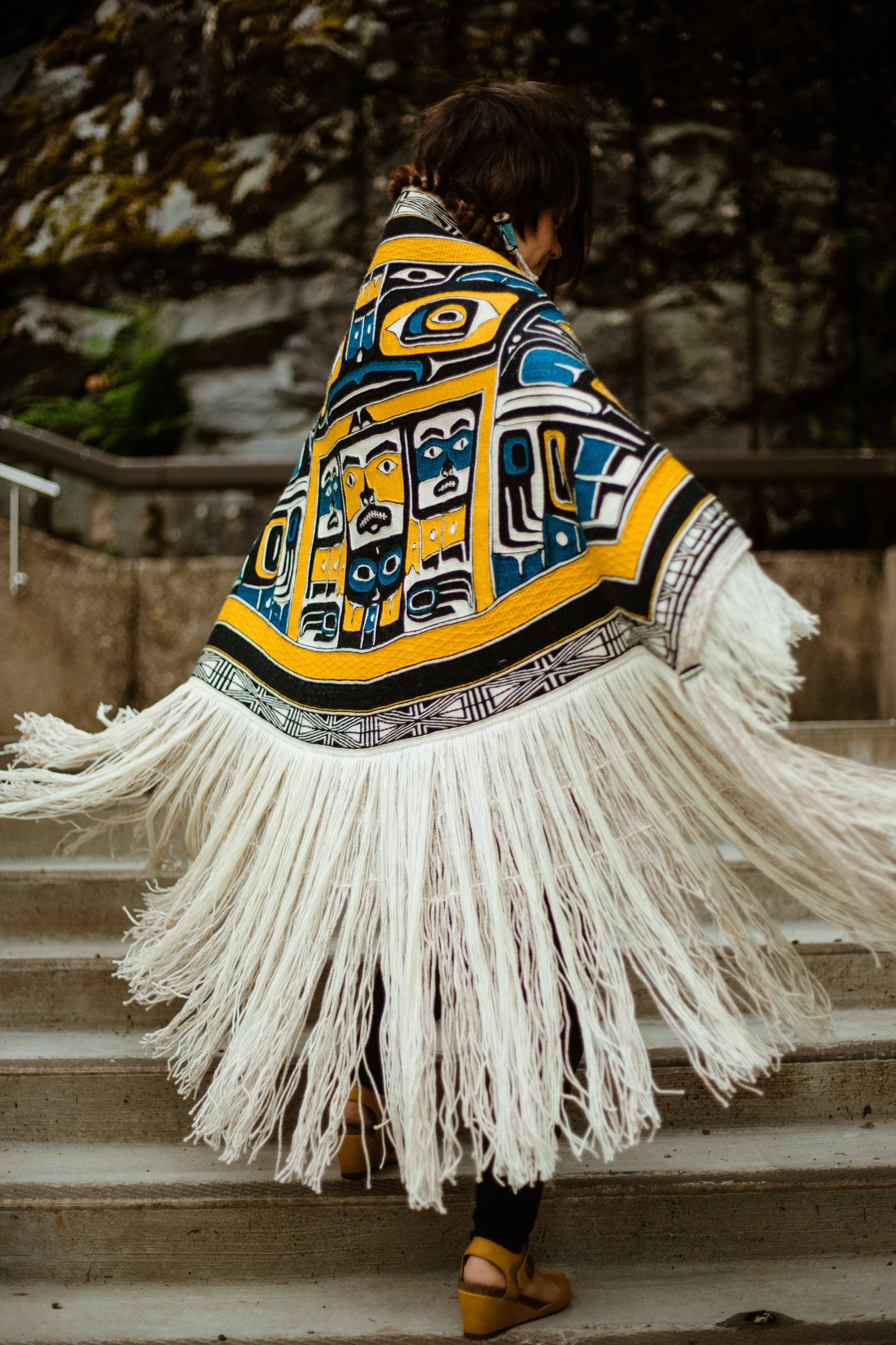 A person wearing a traditional Tlingit Chilkat robe with intricate patterns and long fringe, walking up steps outdoors.