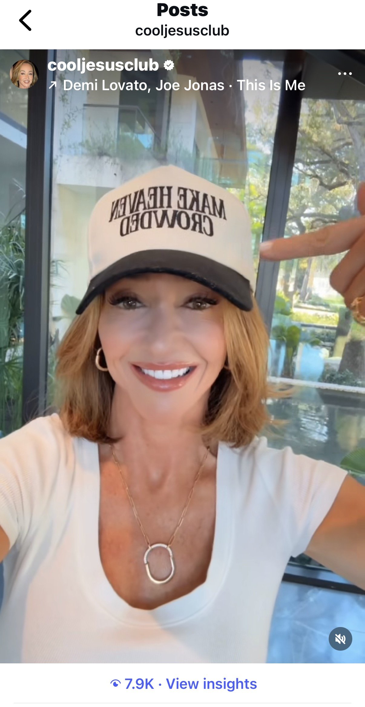 Woman taking a selfie, wearing a white t-shirt, gold necklace with a ring pendant, and a white and black baseball cap that reads 'MAKE HEAVEN CROWDED'.