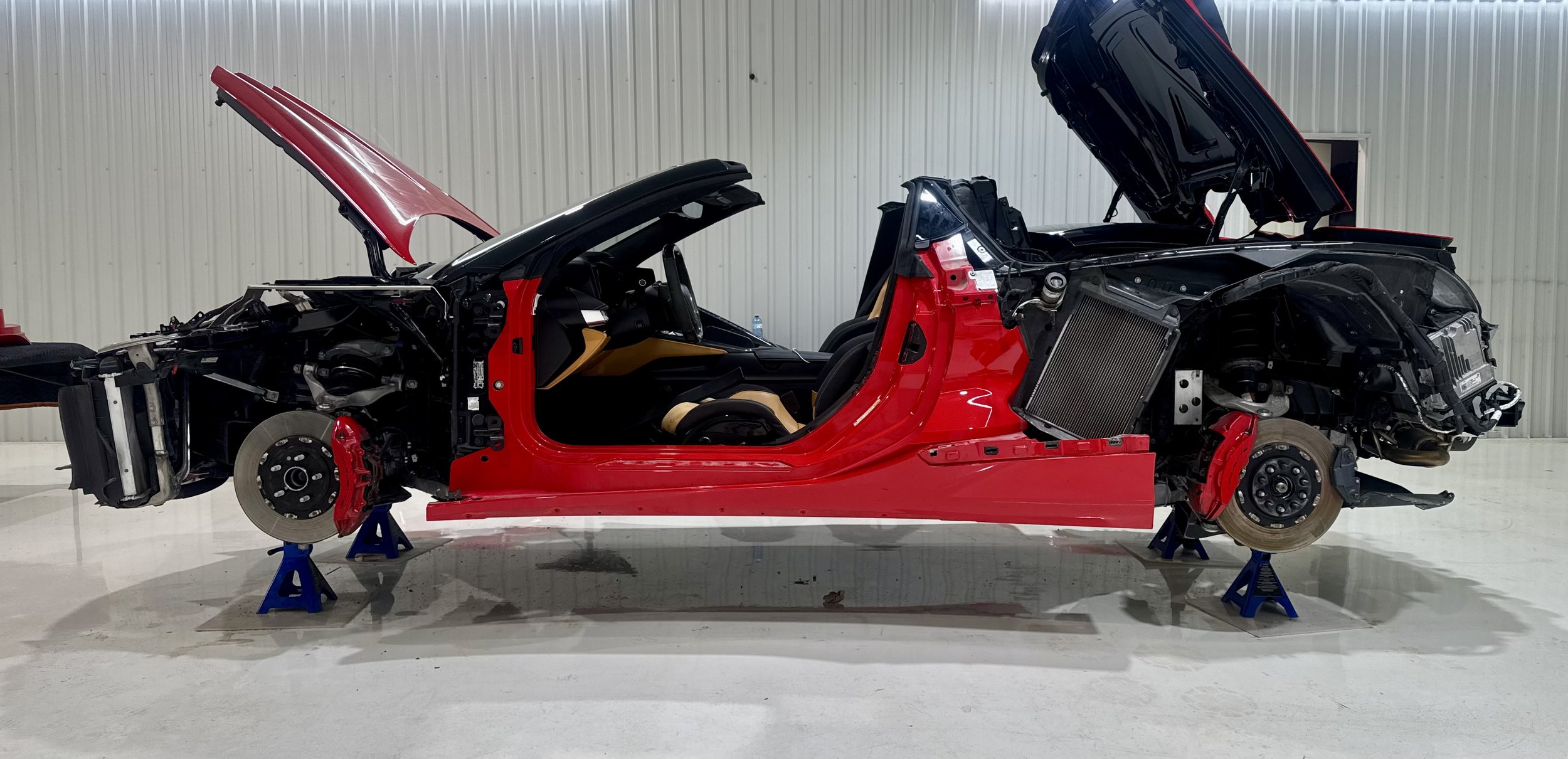 Disassembled red C8 Corvette with no wheels, car lifts under the chassis, and hood open, inside a garage awaiting wrap install.