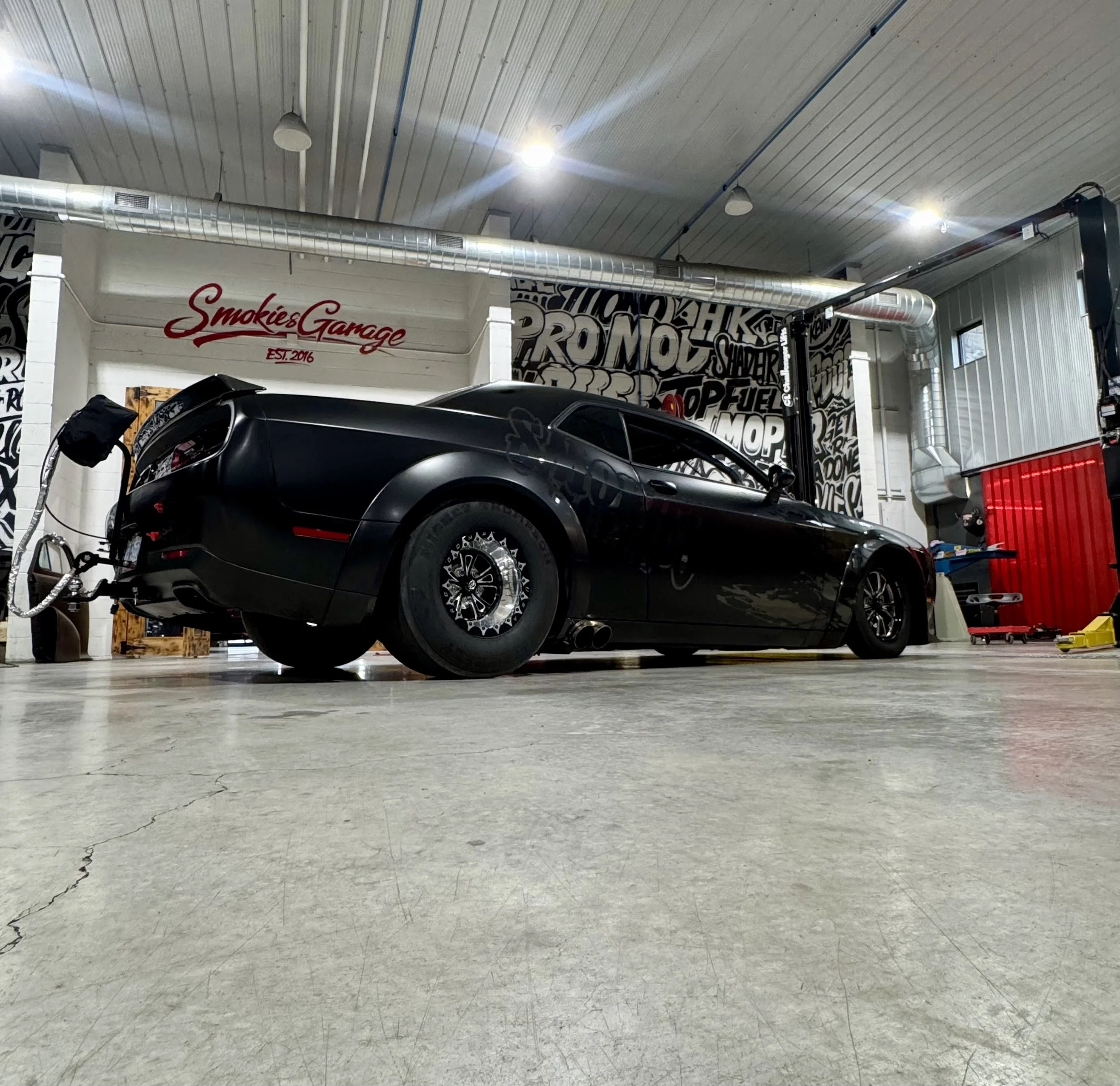 A black race car inside a garage with graffiti-style wall art and a sign that says 'Smokie's Garage'. The car appears to be a drag racing vehicle.