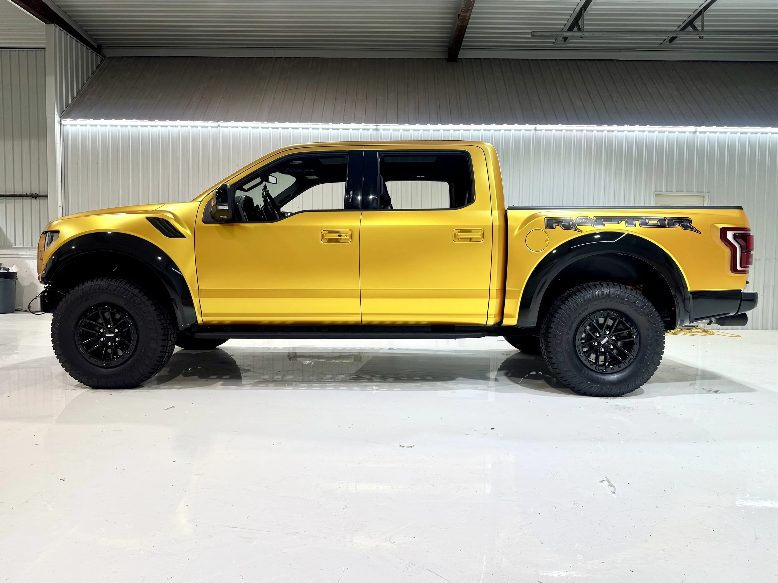 Side view of a wrapped Satin Energetic Yellow Ford Raptor pickup truck inside a warehouse with white walls and a metal ceiling.