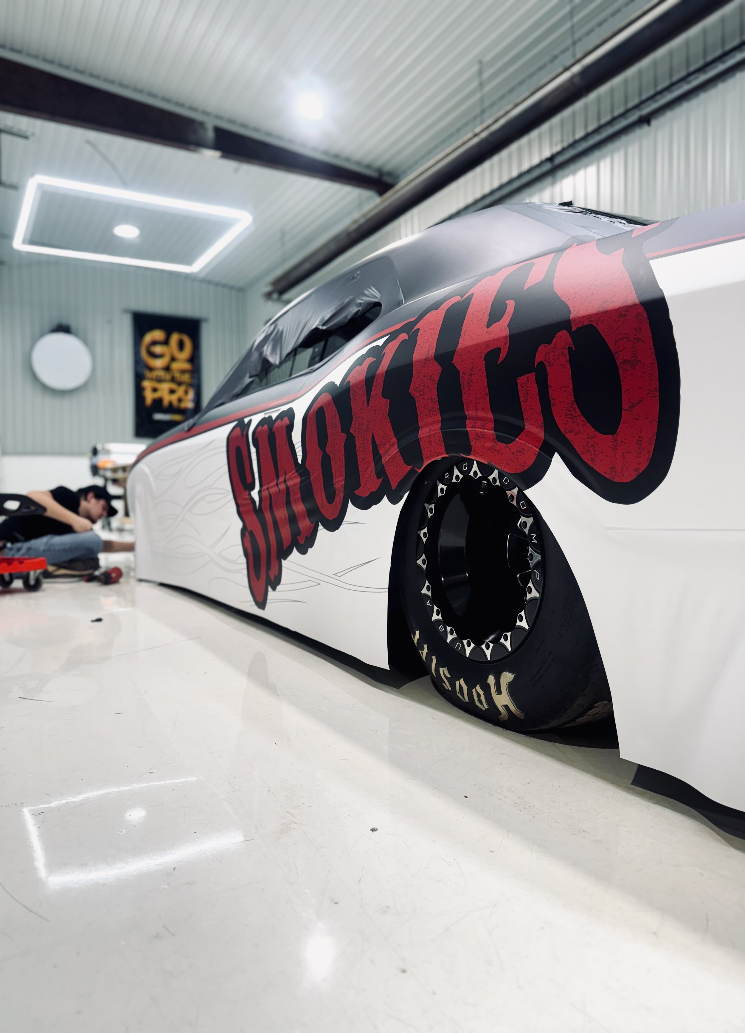 A race car with a large red and black 'SMOKIES' logo on the side, lowered to the ground with a wide wheel and tire setup, inside a well-lit garage with a wrap installer working on the vehicle in the background.