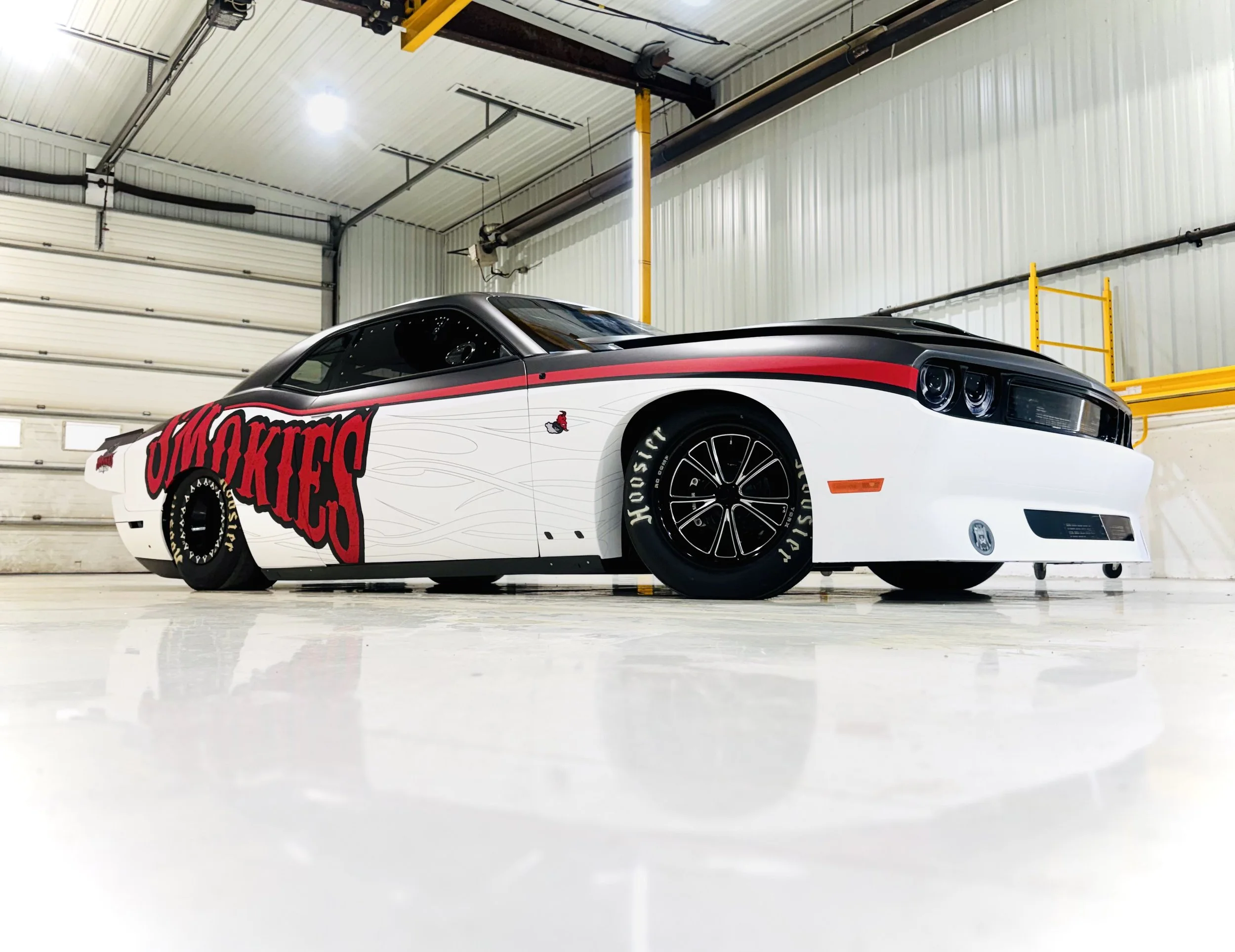 A custom white race car with black accents and red graphics that include the words "SMOKIES" on the side, parked inside a garage with white walls and yellow support beams.