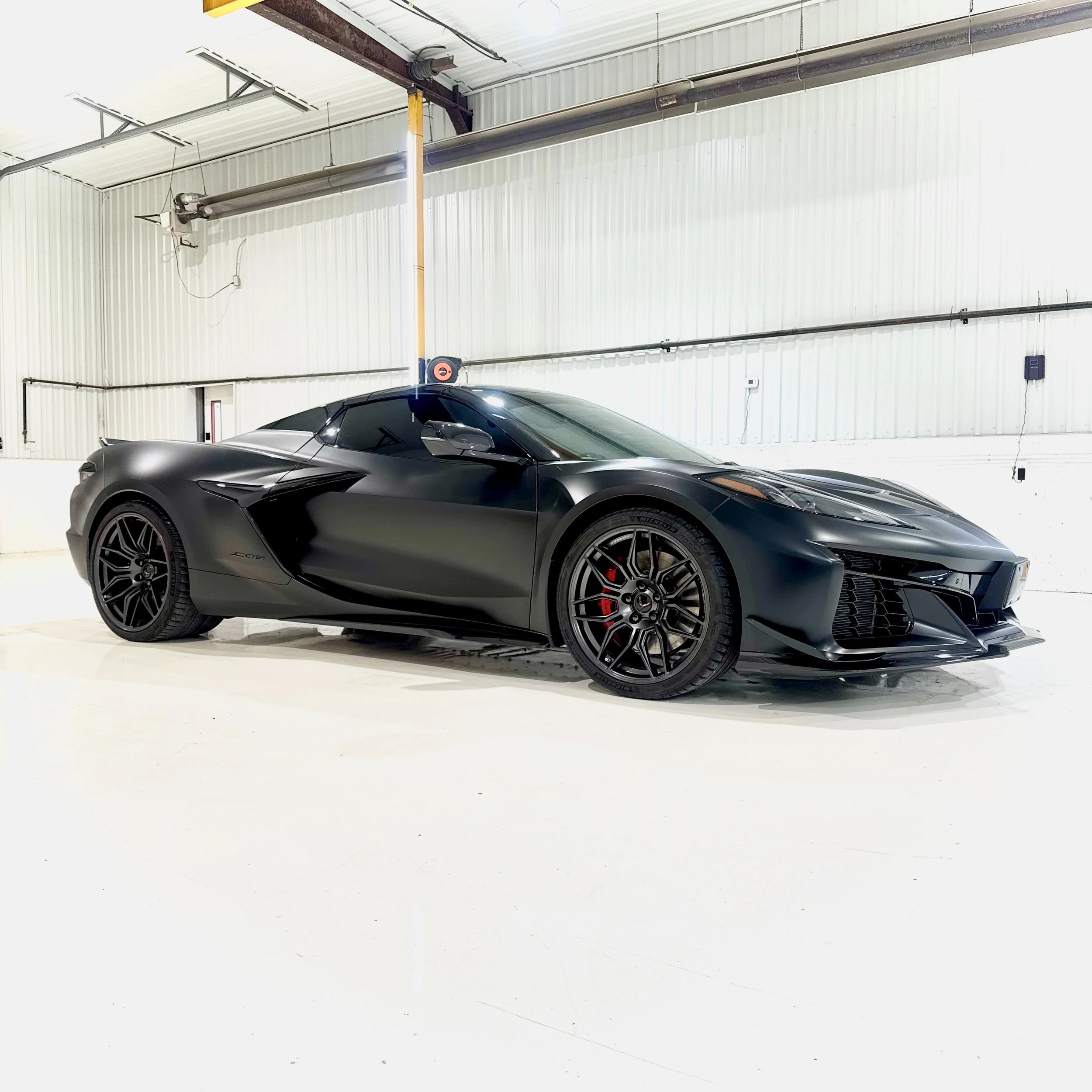 Satin Black wrapped C8 Corvette parked inside a garage with white walls and a high ceiling.