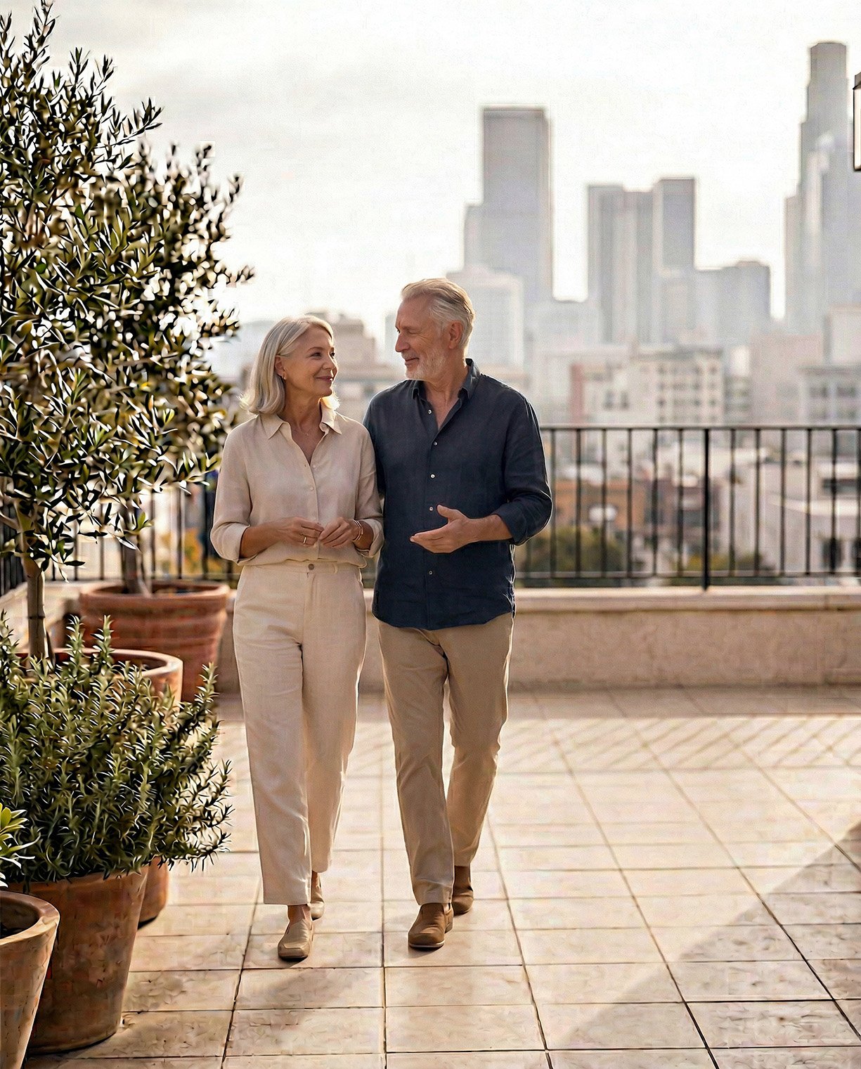 An elderly couple walking and talking on a rooftop patio with city buildings in the background, potted plants nearby.