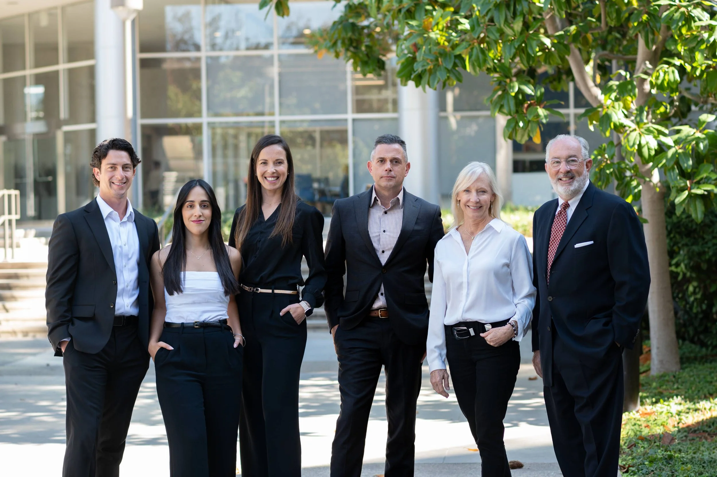 Group of six diverse business professionals standing outdoors in front of a modern office building, smiling at the camera.