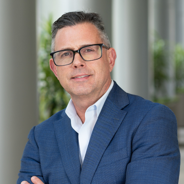 A man wearing glasses in a blue suit and white shirt, posing indoors with a blurred background of greenery and natural light.