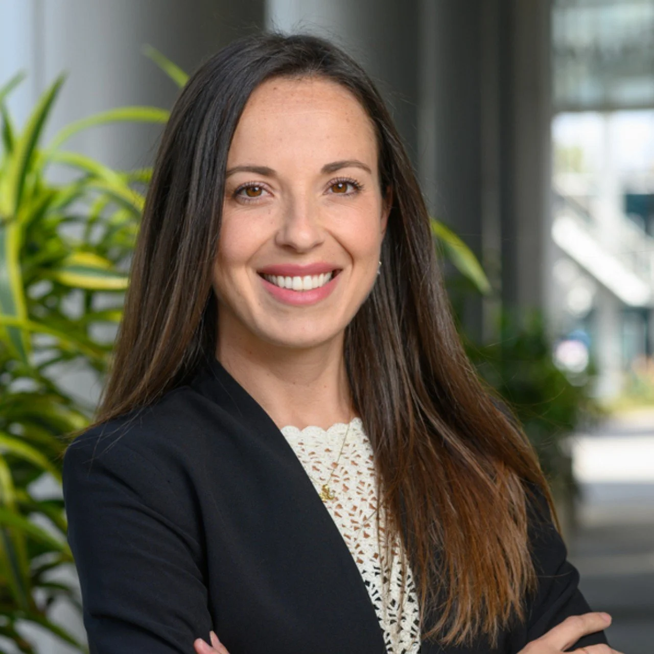 Portrait of a smiling woman with long brown hair, wearing a black blazer and white lace top, standing indoors with green plants and a modern building background.