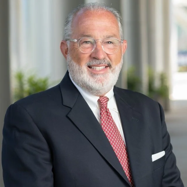 Portrait of a smiling elderly man with gray hair and a beard, wearing a dark suit, white shirt, and red patterned tie, indoors with blurred background.