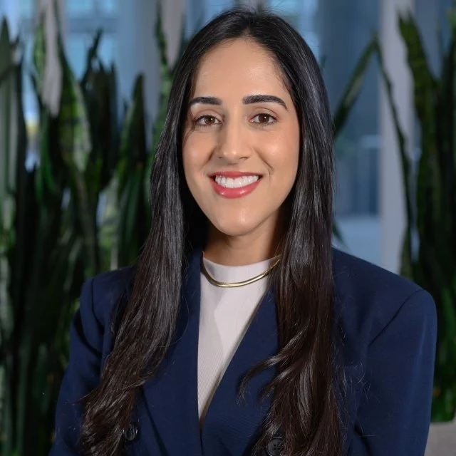A young woman with long dark hair, wearing a navy blazer and a gold necklace, smiling confidently in an office setting with green plants in the background.
