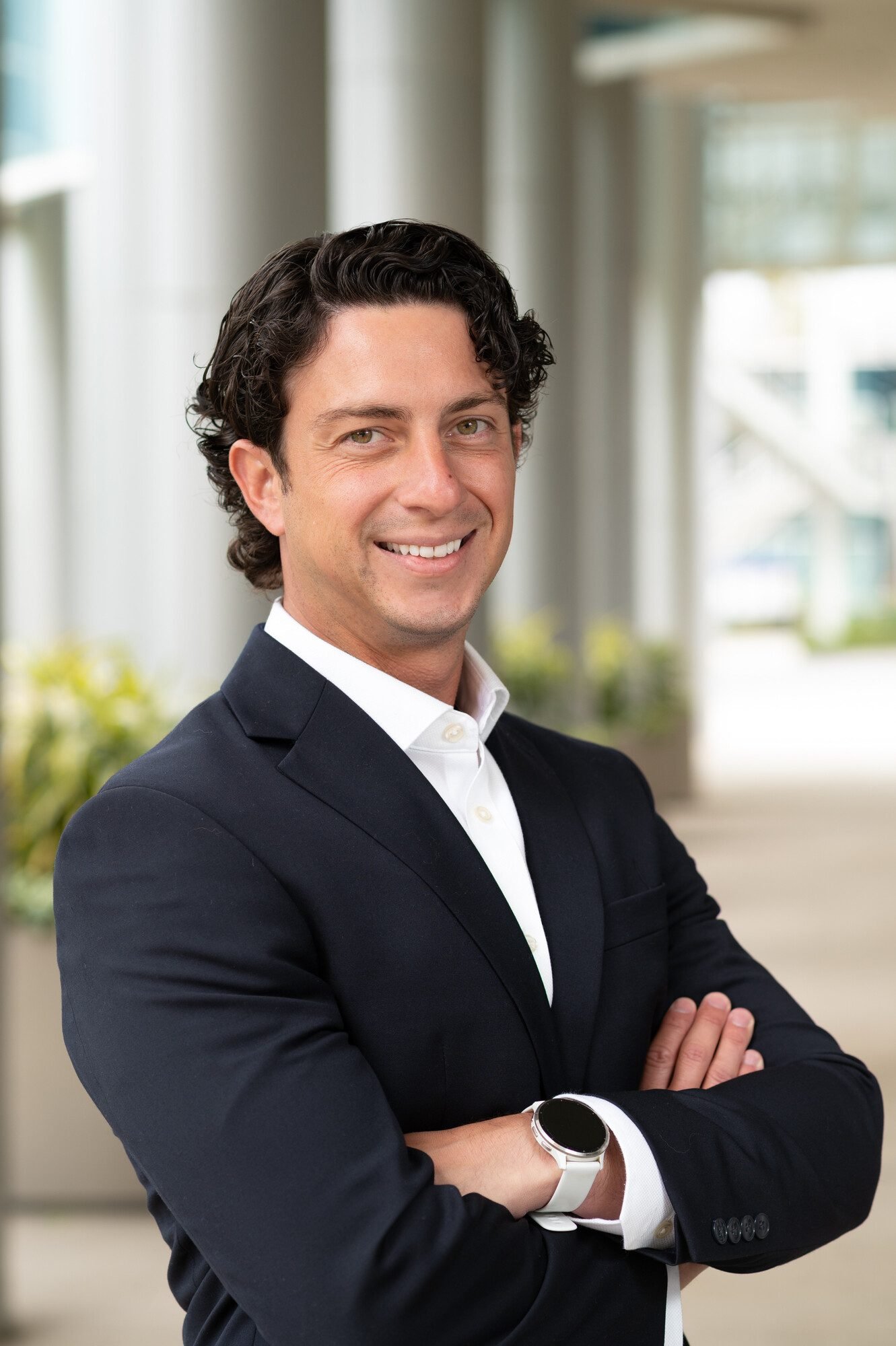 A smiling man with dark curly hair standing with arms crossed, wearing a dark suit, white shirt, and a smart watch, in a bright office environment with windows and plants in the background.
