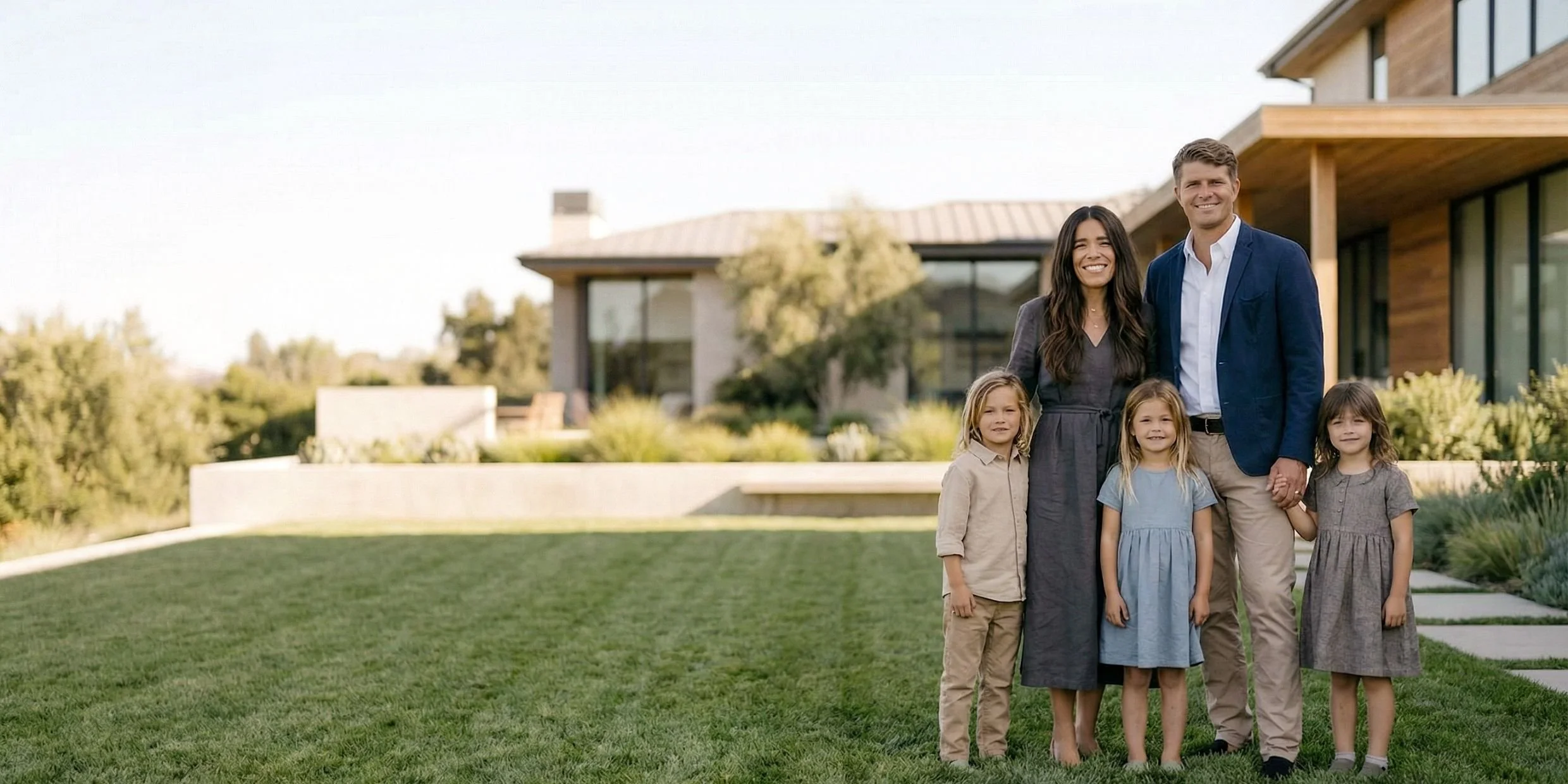 A family of five standing outside a modern house with a lawn. Two adults, a woman and a man, are holding hands, with three young girls standing beside them. All are smiling and dressed casually.