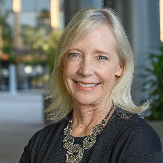 A smiling middle-aged woman with blonde hair, wearing a black top and a large spiral-patterned necklace, standing indoors with a window view of a cityscape in the background.