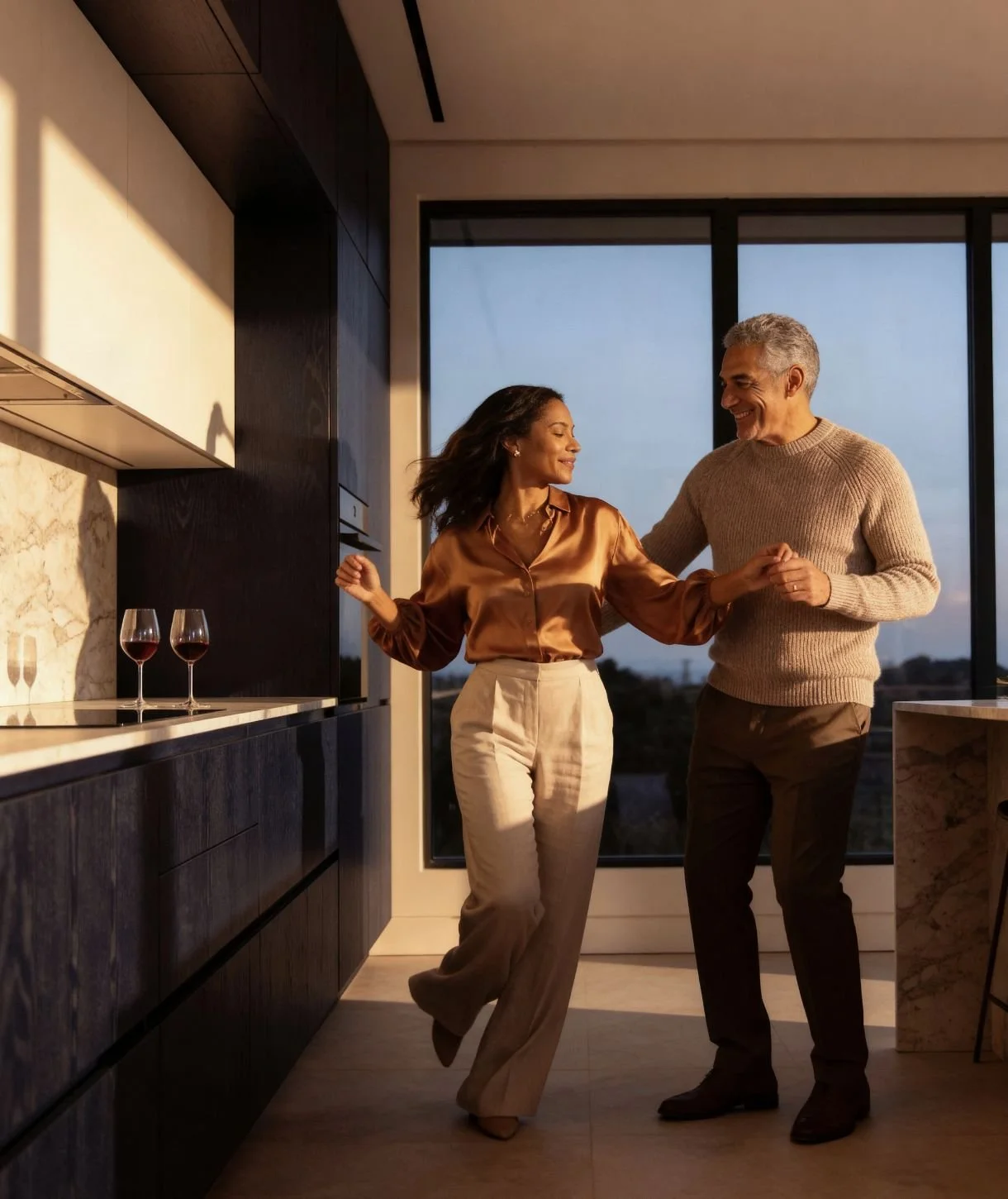 A couple dancing in a modern kitchen with large windows showing an evening sky. Two glasses of red wine are on the counter.