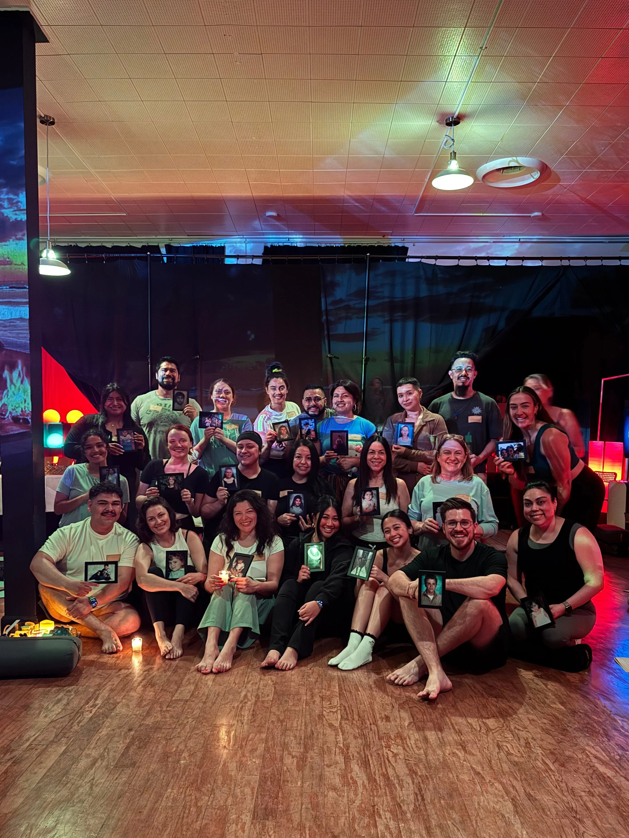 A diverse group of people gathered indoors, sitting and standing on a wooden floor, holding framed photographs, with dim lighting and colorful stage lights in the background.