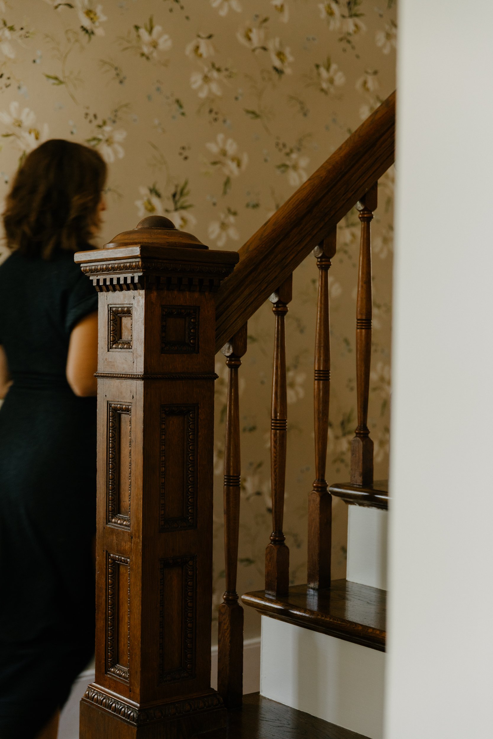 A woman with brown hair wearing a black top walking past a wooden staircase with decorative carved details, beige floral wallpaper on the wall behind.