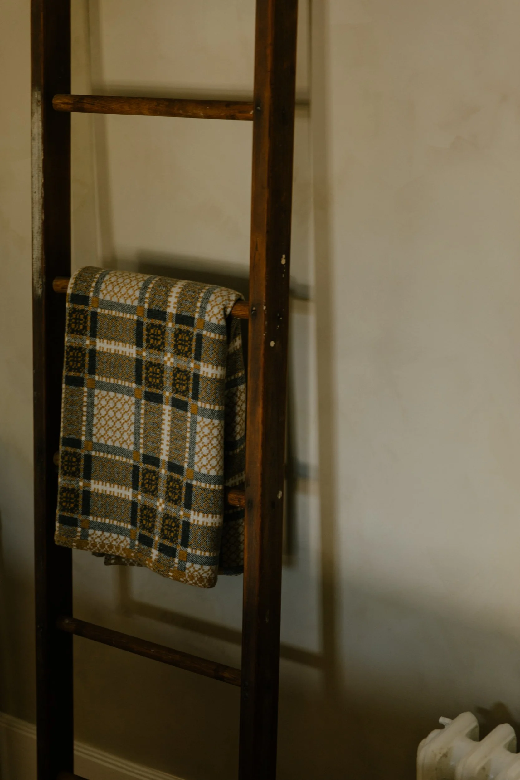 A wooden towel rack with a patterned towel hanging on it, positioned against a beige wall in a room with a heating radiator visible at the bottom right corner.