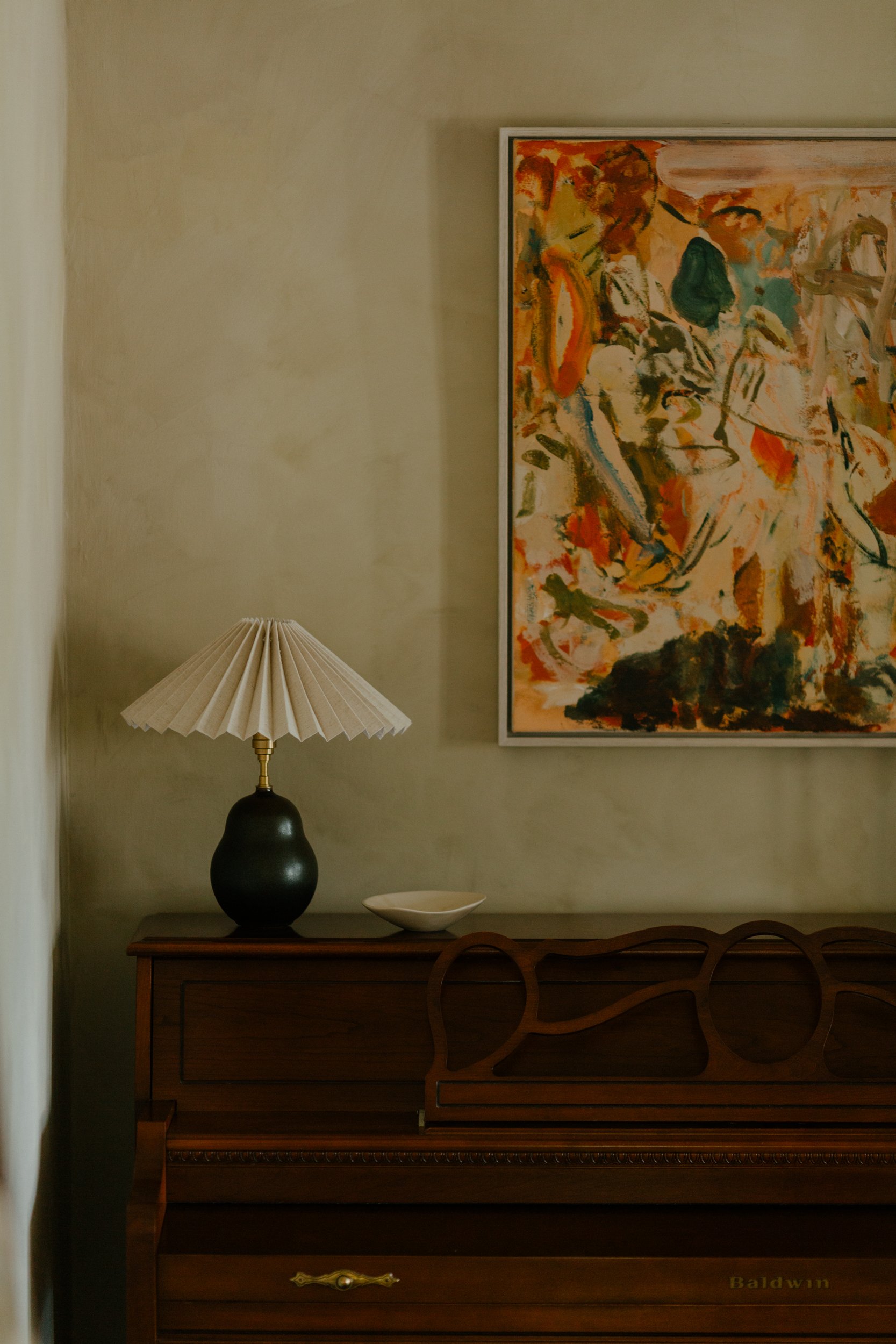 A corner of a room with a beige wall, a colorful abstract painting hanging, and a dark wooden piano with a brass handle, a white ceramic bowl, and a tall black lamp with a pleated beige lampshade.