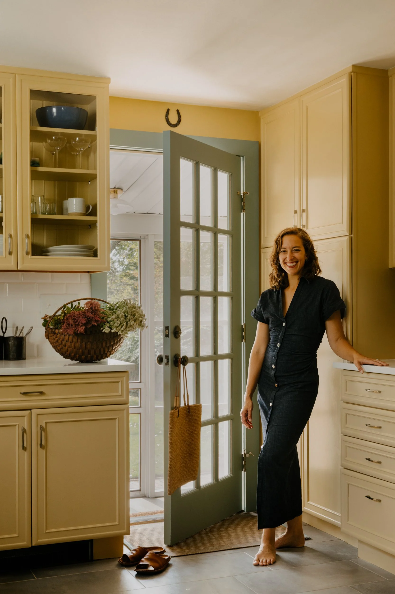 A woman standing barefoot in a beige kitchen near an open door, smiling at the camera. There are beige cabinets, a basket of flowers on the counter, a pair of brown slippers on the floor, and a horseshoe hanging above the door.