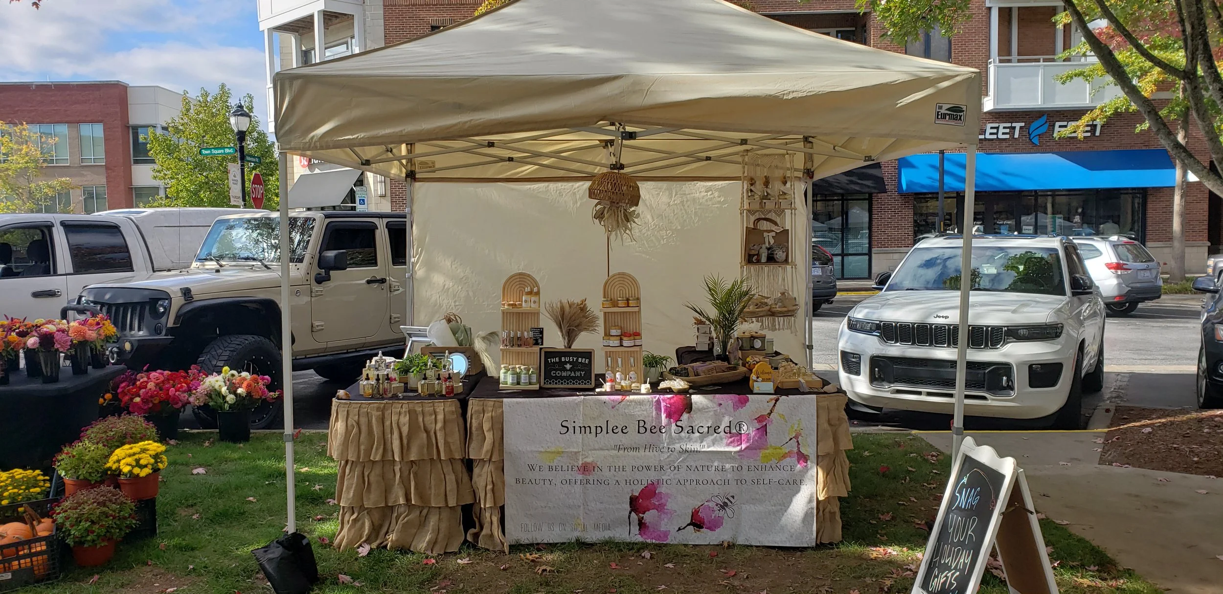 Market stall named 'Simplee Bee Sacred' selling natural beauty products and self-care items under a tent in an outdoor setting, with flowers on either side and parked cars in the background.