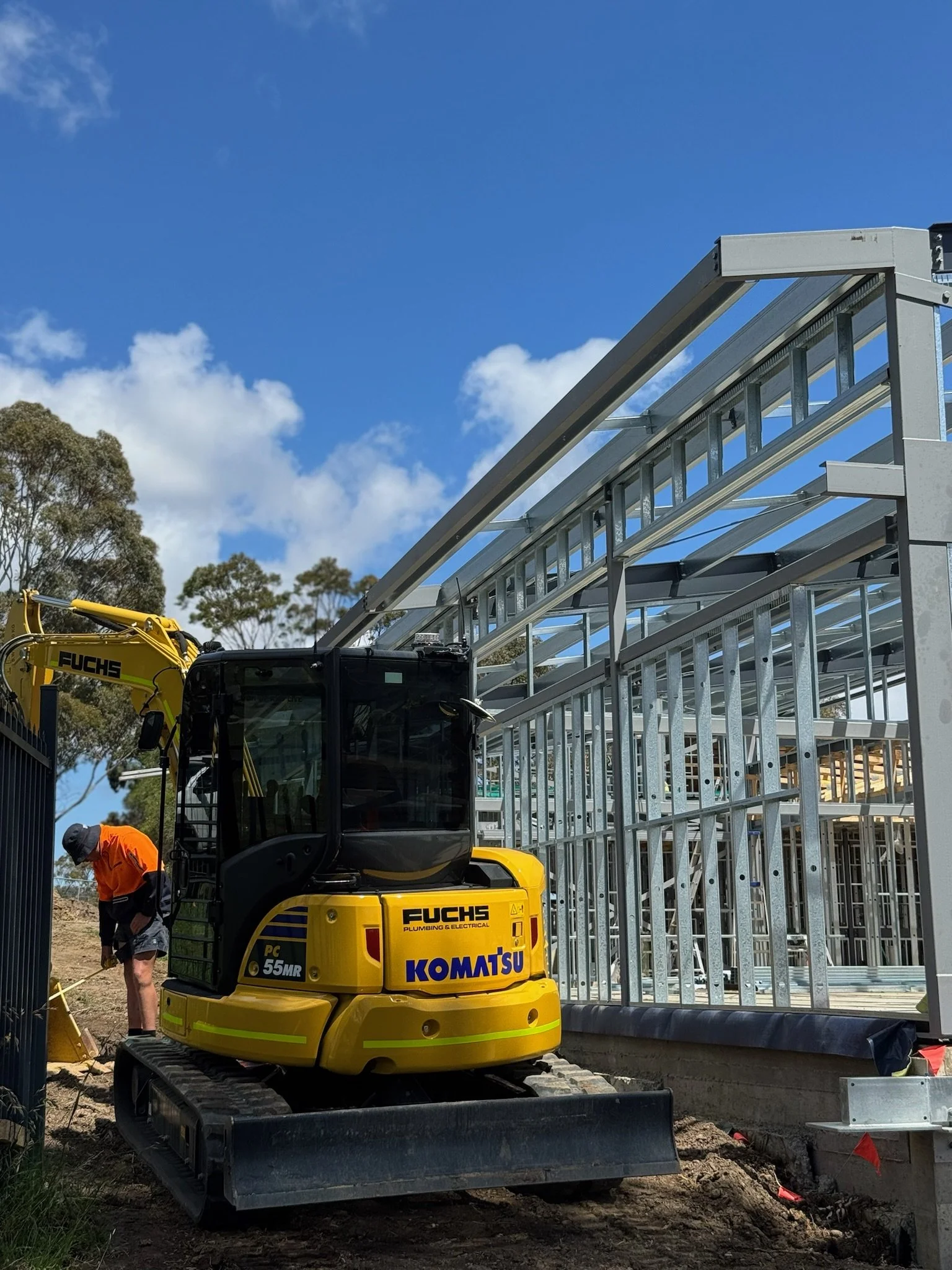Construction site with a yellow Komatsu mini excavator and a worker in an orange shirt and helmet working near a metal frame structure under a blue sky with scattered clouds.