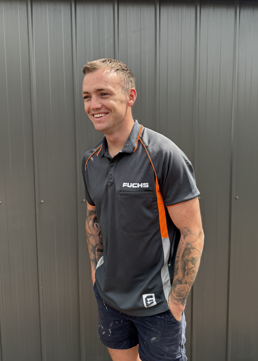 A smiling man with tattoos on his arms wearing a black and orange Fuchs sports shirt and dark shorts, standing in front of a gray metal wall.