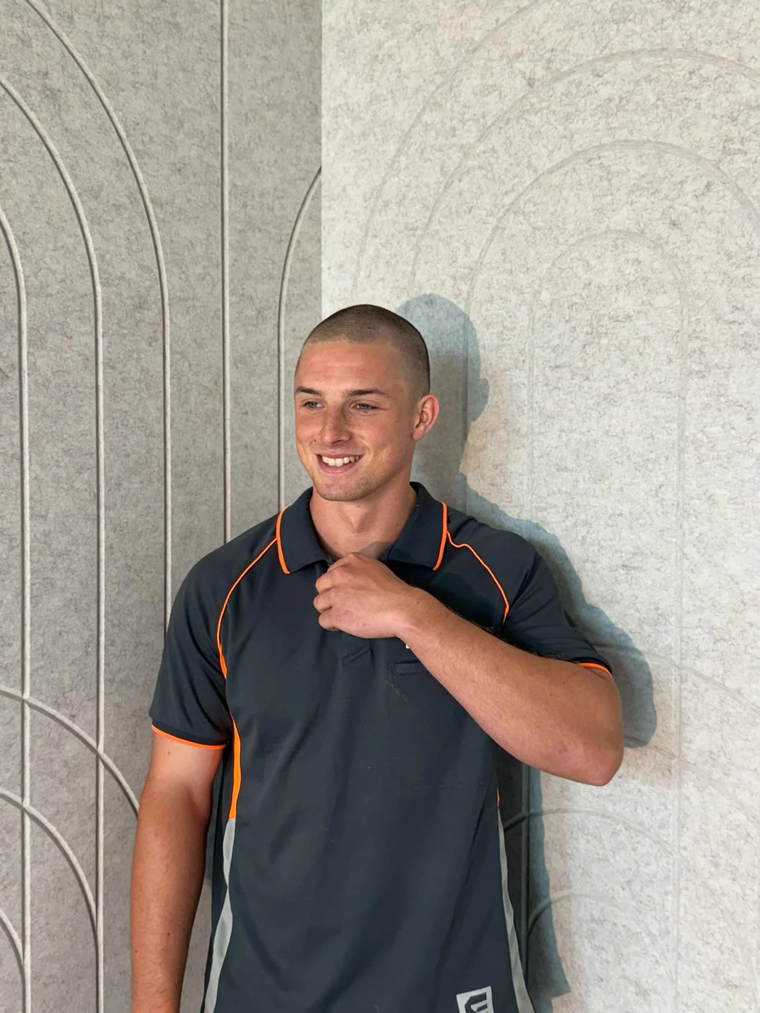 A young man with a shaved head smiling and standing against a curved gray wall, wearing a black sports shirt with orange accents.
