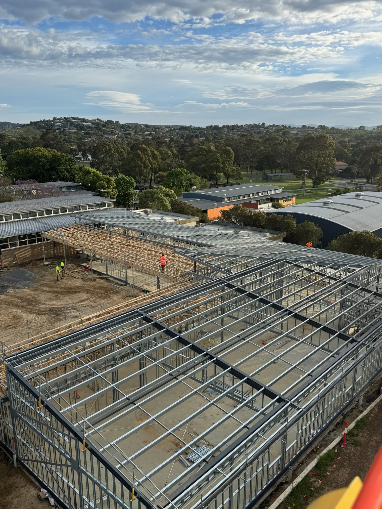 Construction site with steel framework for a building, workers in high-visibility clothing working on the structure, and trees and buildings in the background under a partly cloudy sky.