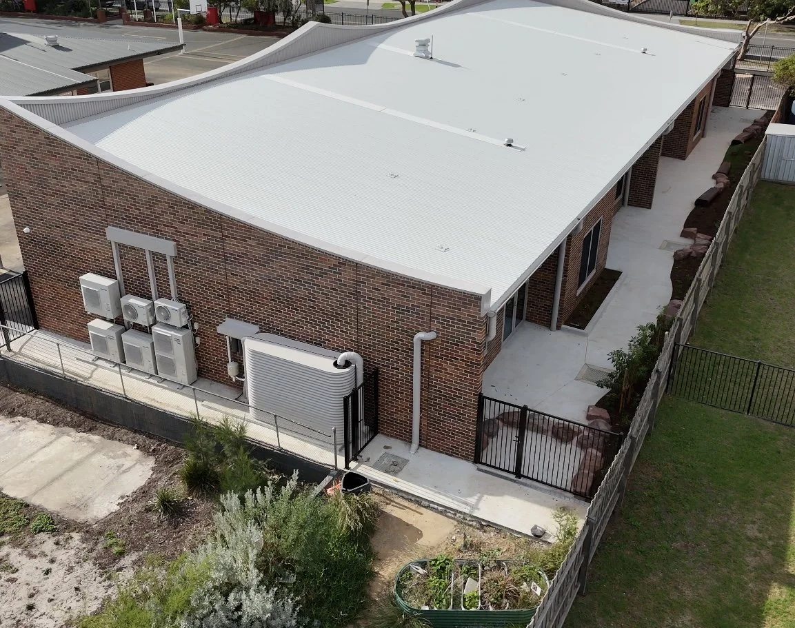 Aerial view of a brick building with a white metal roof, multiple HVAC units on one side, and a fenced backyard with a concrete patio and landscaping.